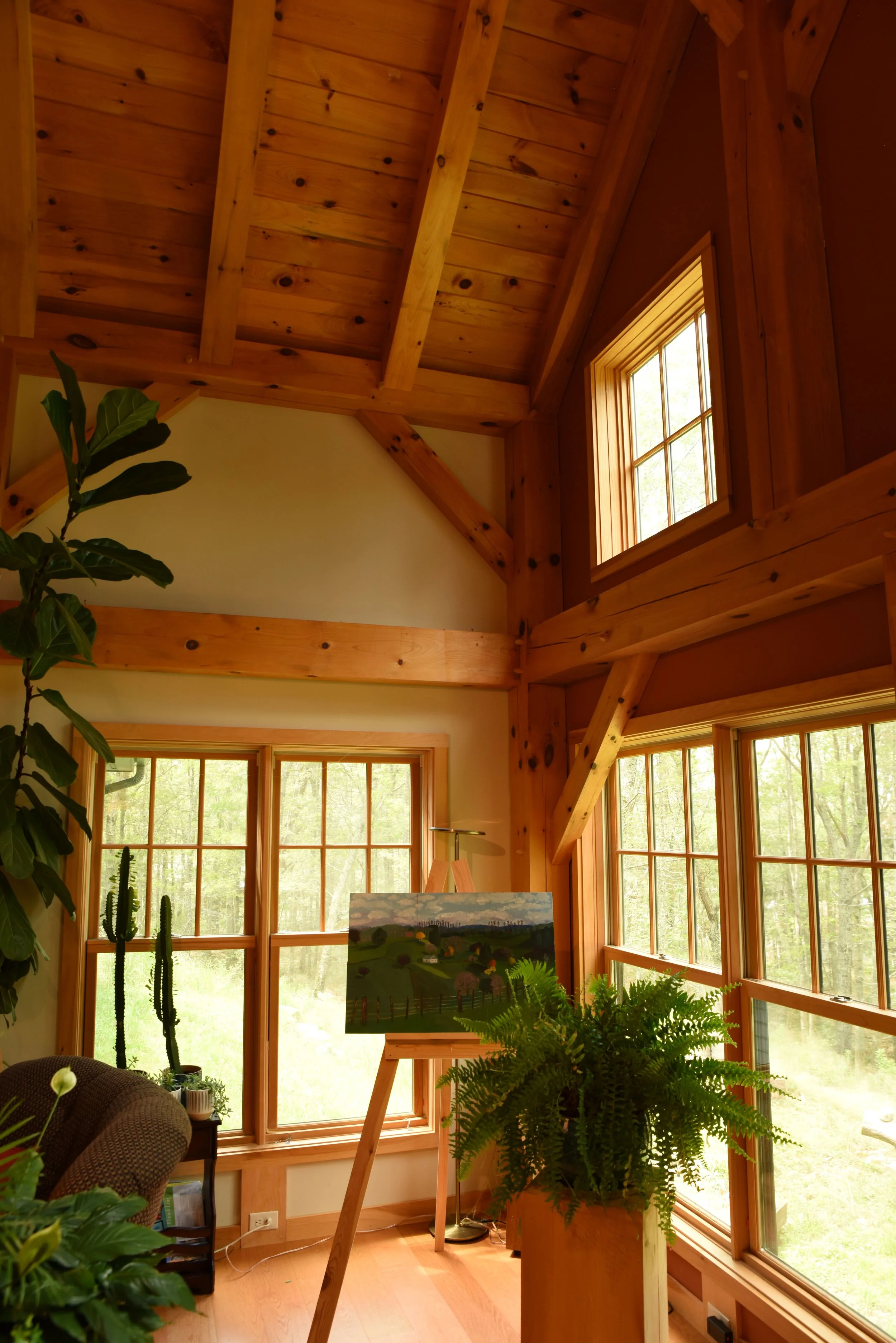 Interior of a wooden room with large windows, a potted fern, an easel with a landscape painting, and a dining chair in front of a window.