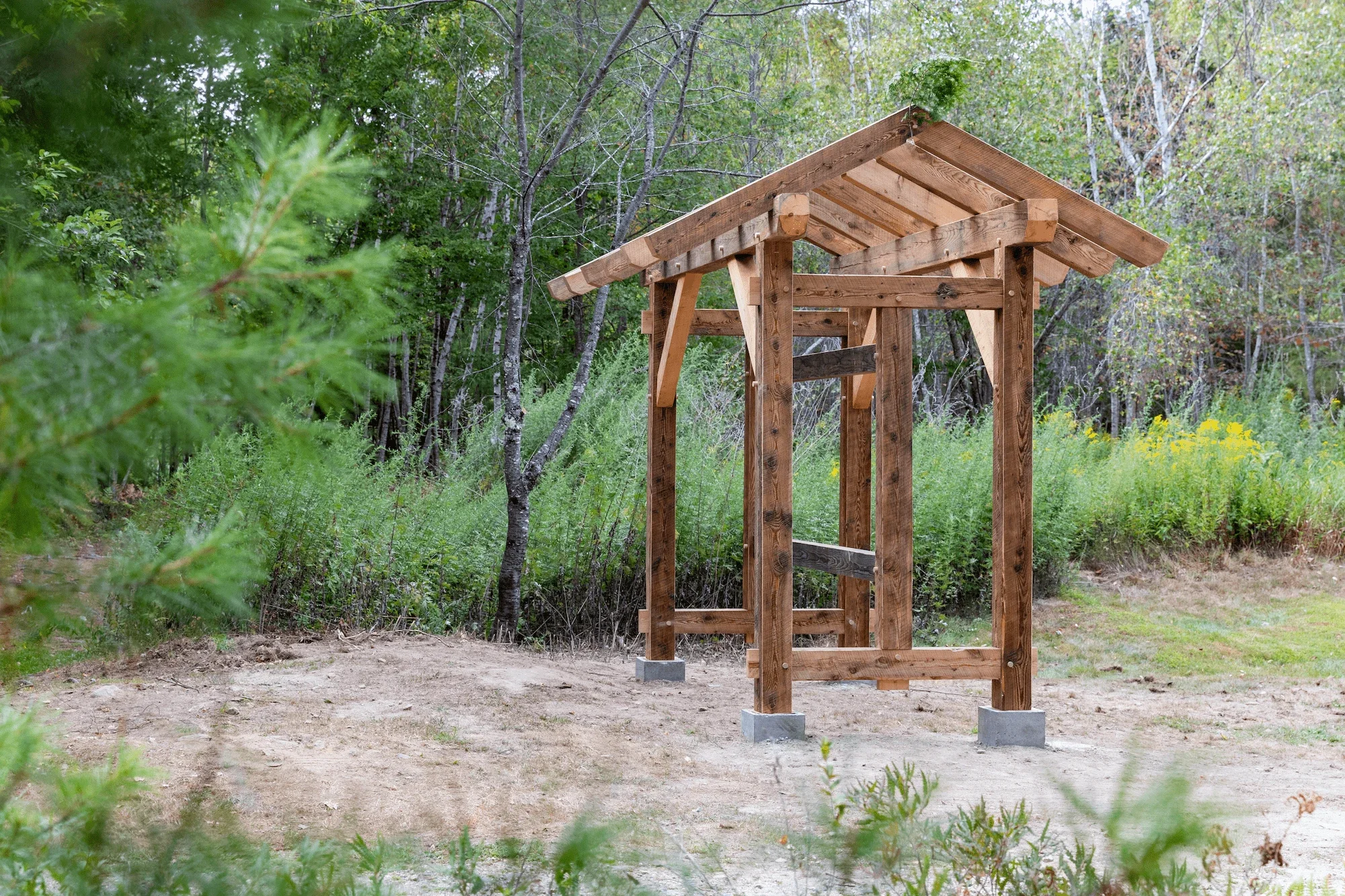 A wooden outdoor structure with a sloped roof, situated in a natural setting with trees and green foliage in the background.