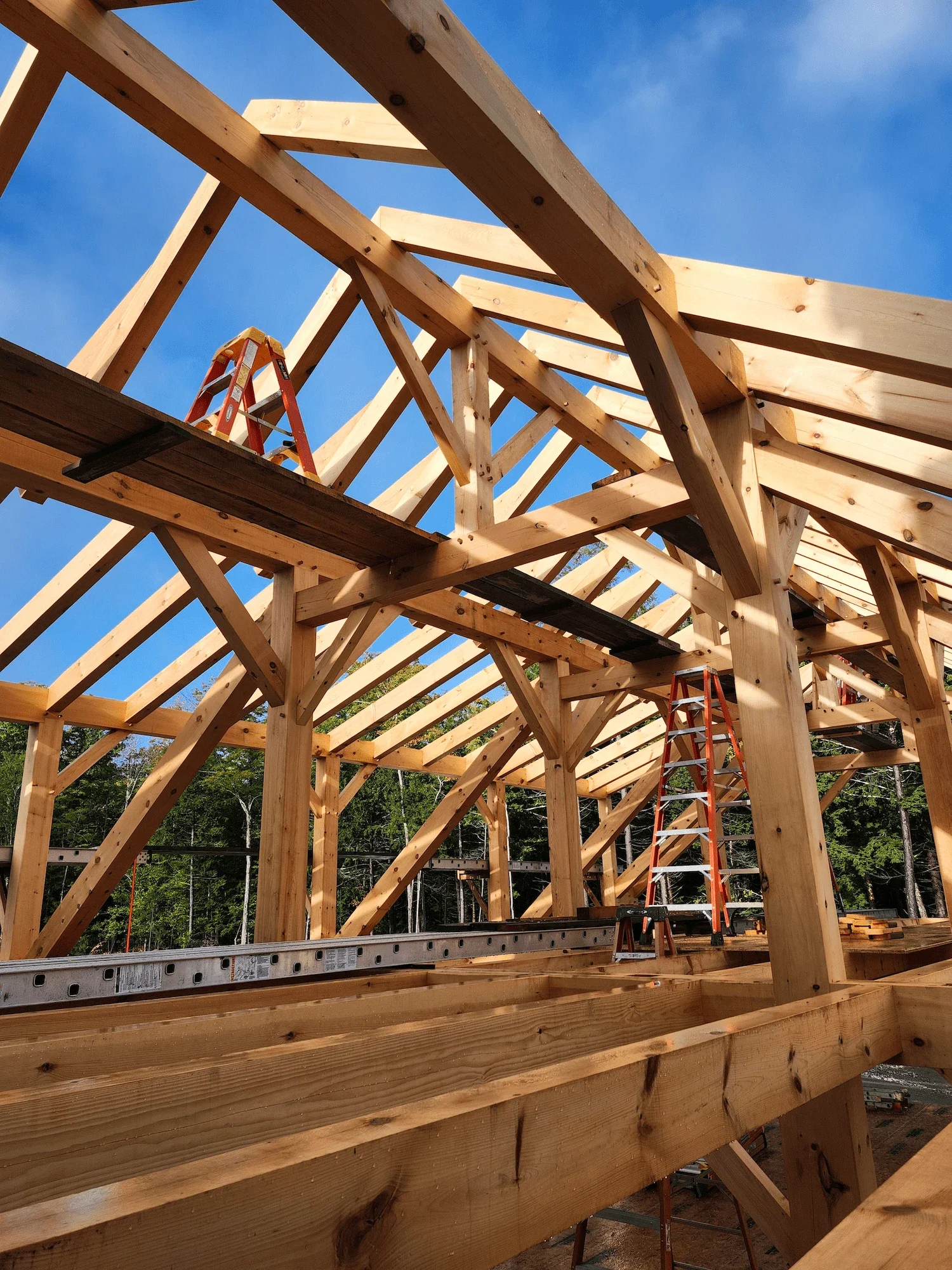 Wooden house framework under construction with ladders, set against a blue sky with some clouds.