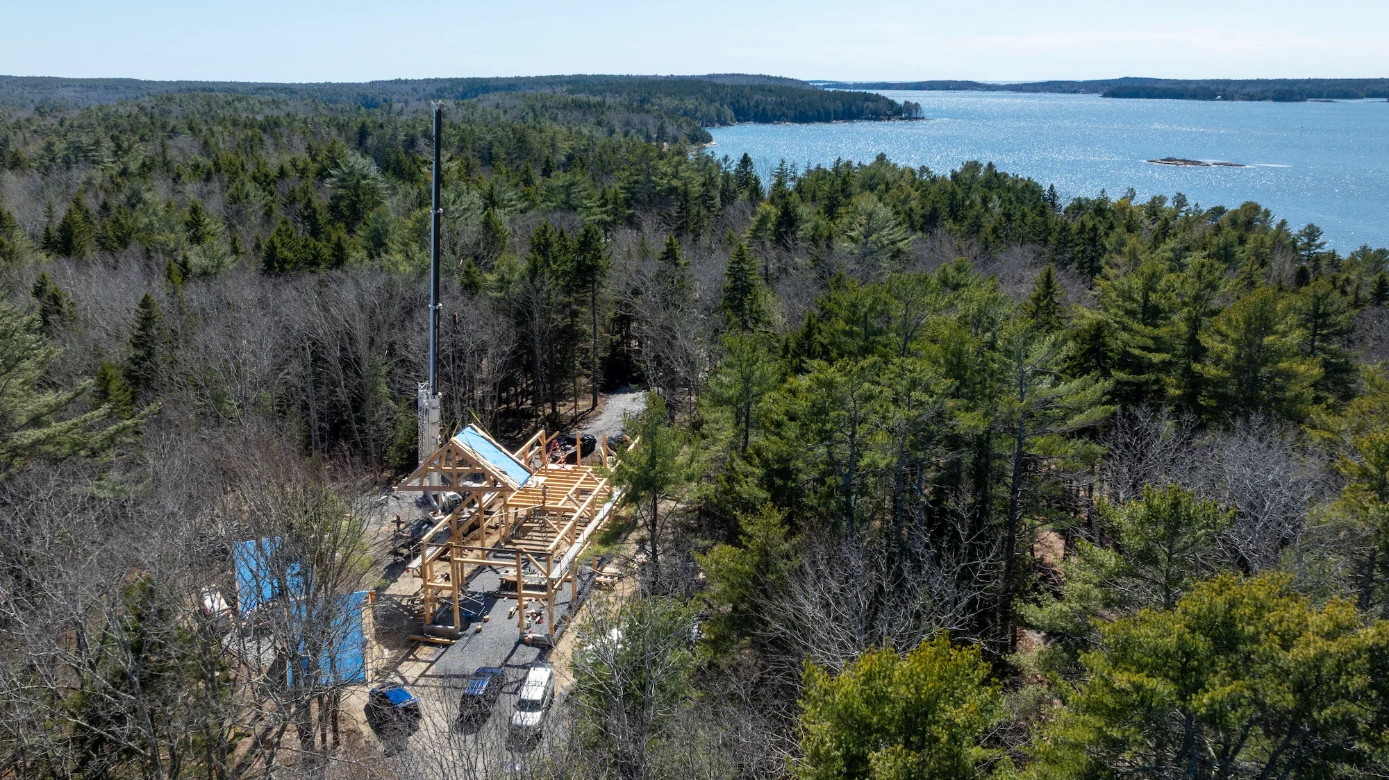 Aerial view of a construction site in a forested area near a lake, with a tall antenna, a partially built wooden structure, and several parked vehicles.