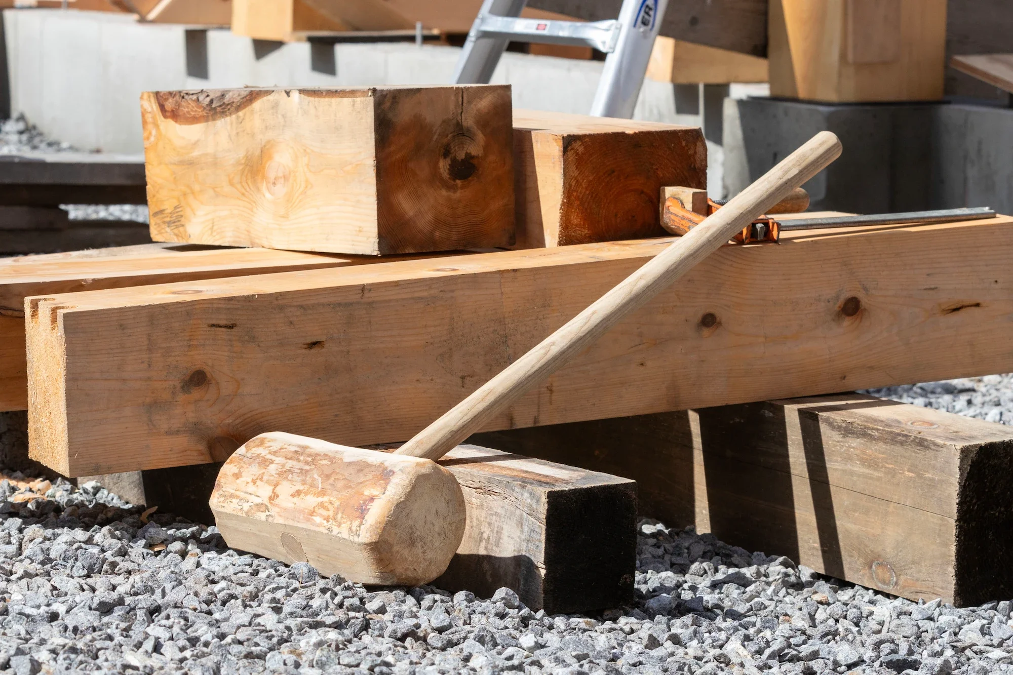 A woodworking site with large wooden beams, a hammer resting on a black block, and a saw resting on a beam, surrounded by gravel.