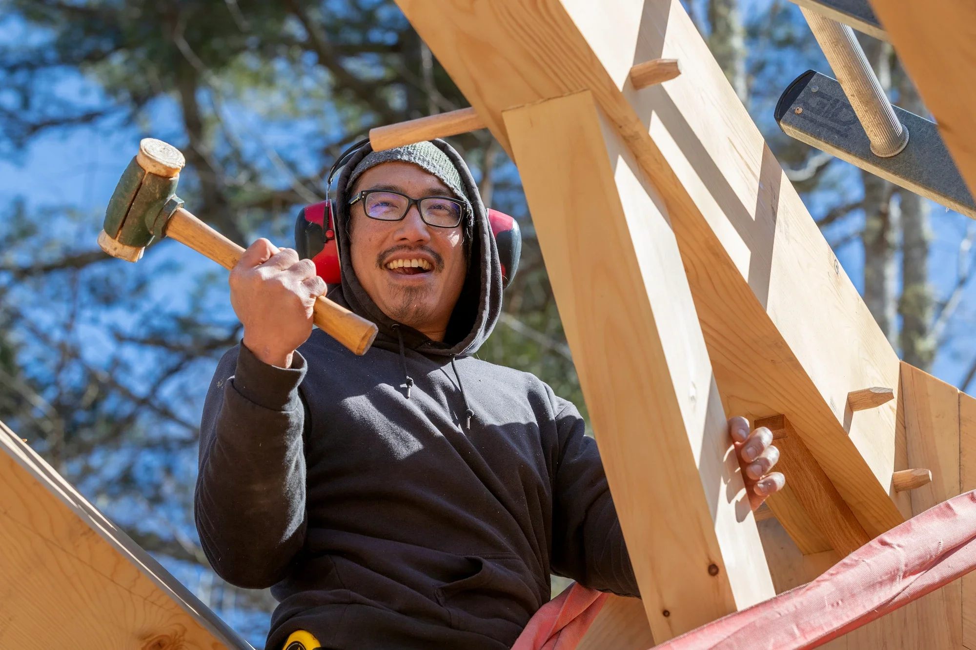 A man wearing glasses, on a winter day, holding a hammer, smiling, and working on a wooden construction outdoors.