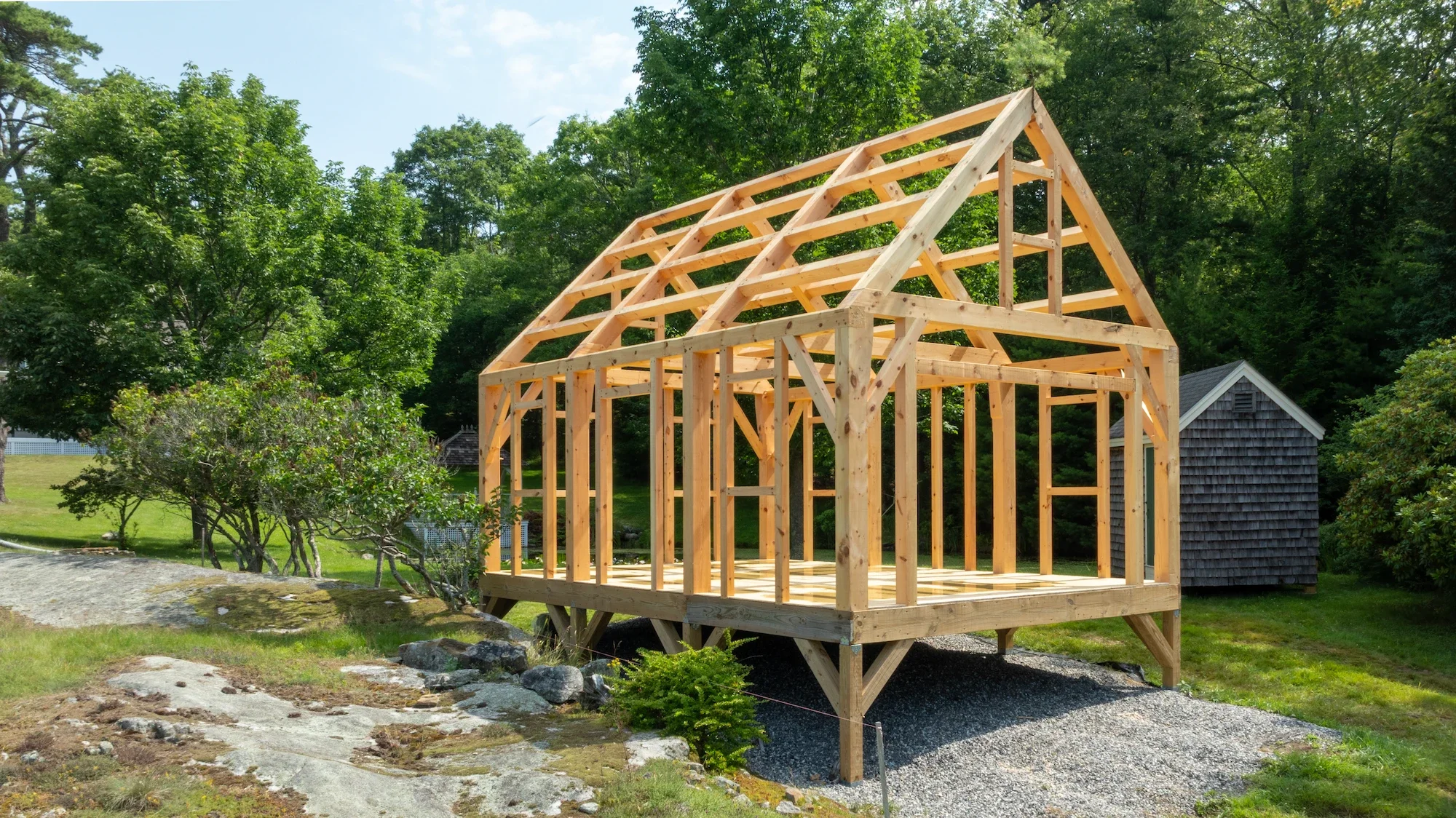 Wooden house frame under construction outdoors with green trees and a small shed in the background.