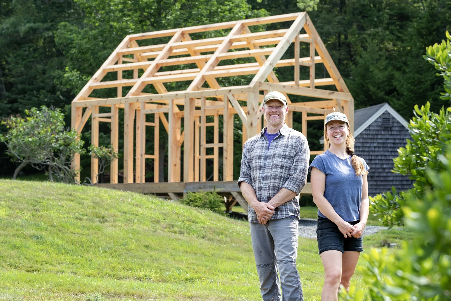 Smiling man and woman outdoors in front of partially built wooden house frame, with lush green trees and a small shed in the background.