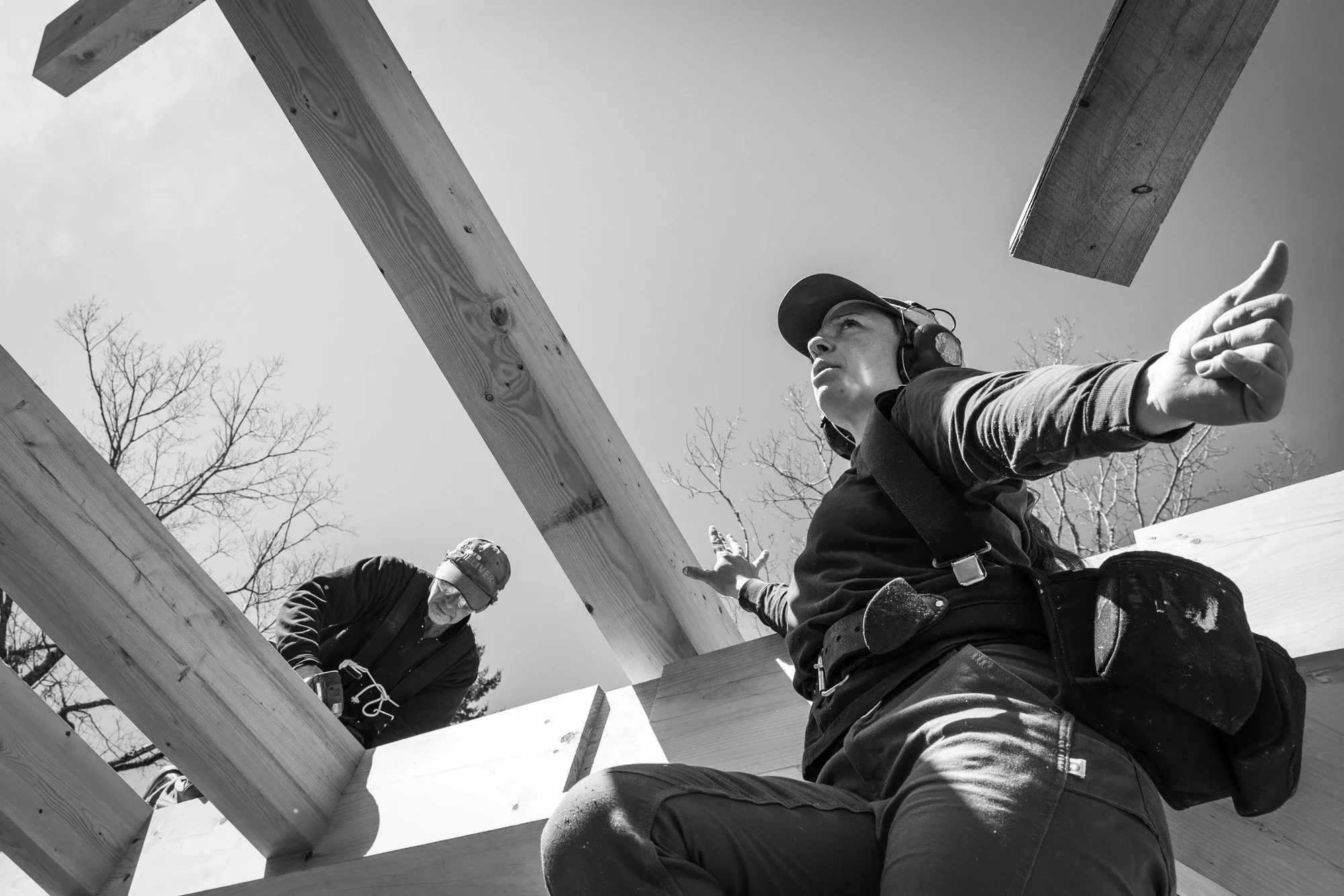 Two construction workers building a wooden structure outdoors, with one worker pointing, both wearing helmets and safety gear, with leafless trees in the background.
