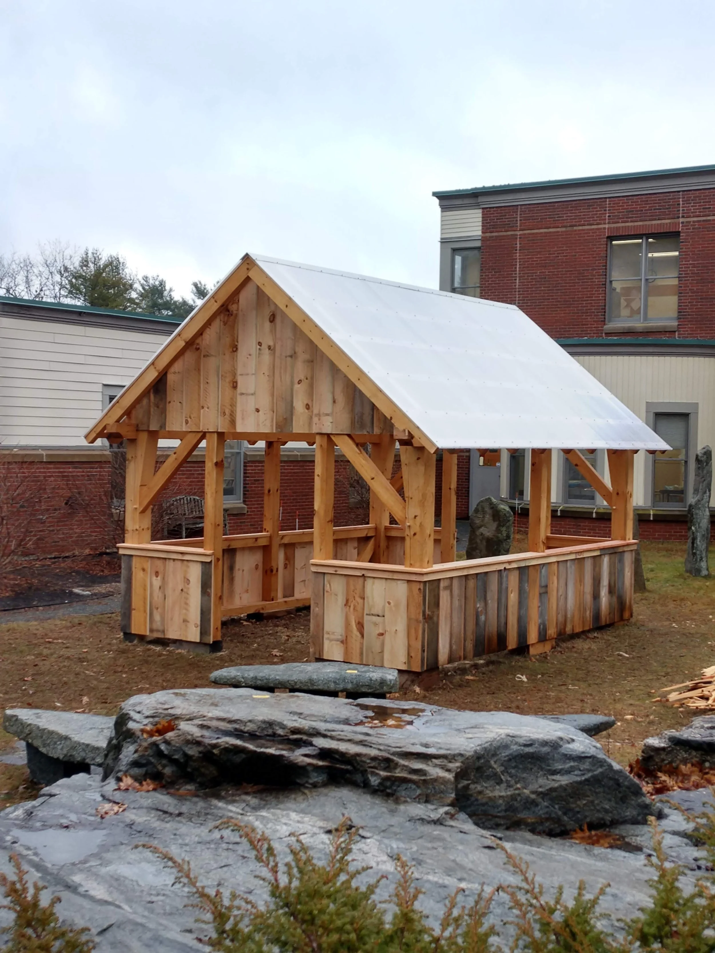Small wooden structure with a pitched roof in a yard, surrounded by rocks and grass, with buildings in the background.