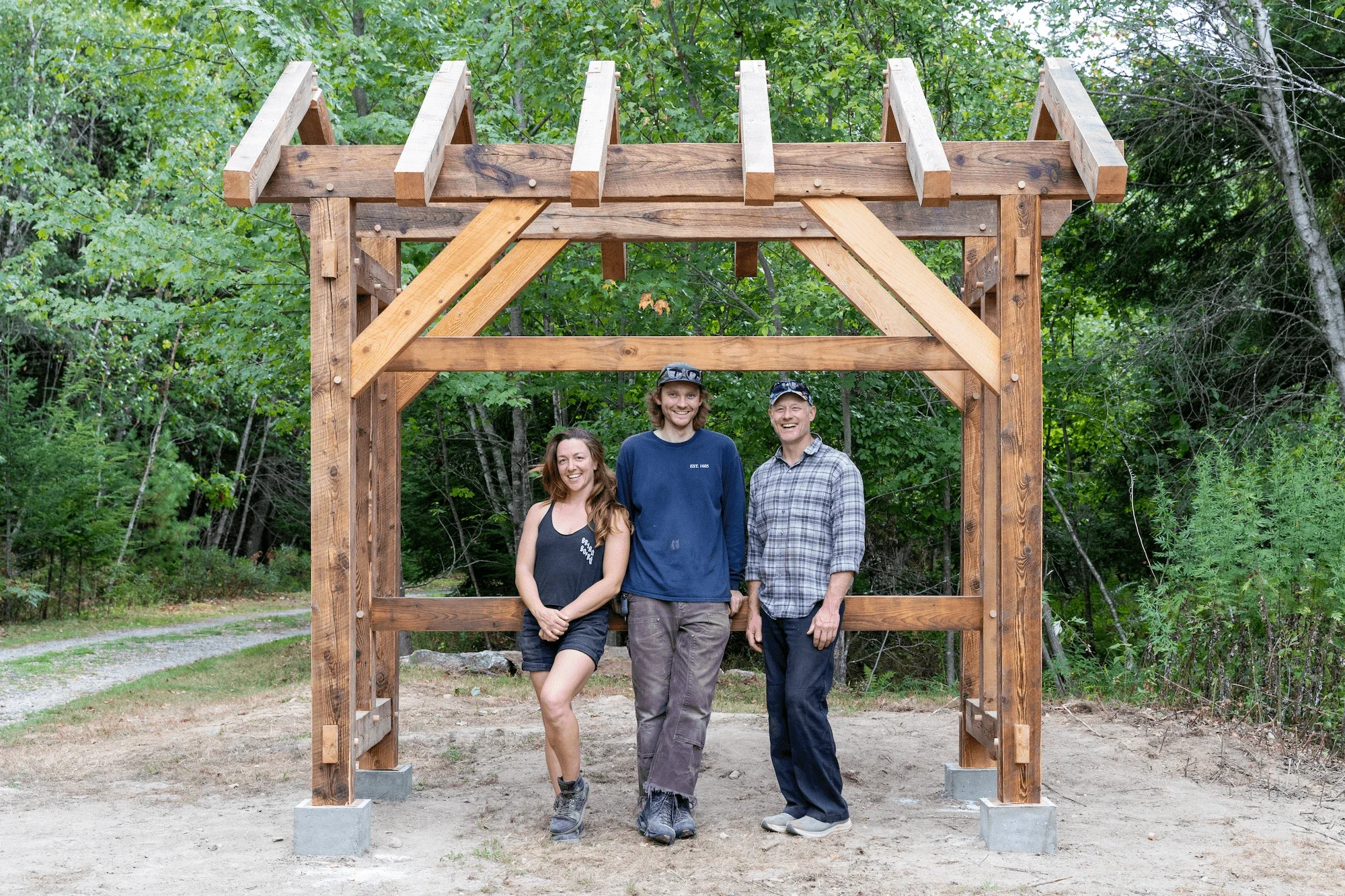 Three smiling people, two men and a woman, standing under a wooden structure frame in a forested area.