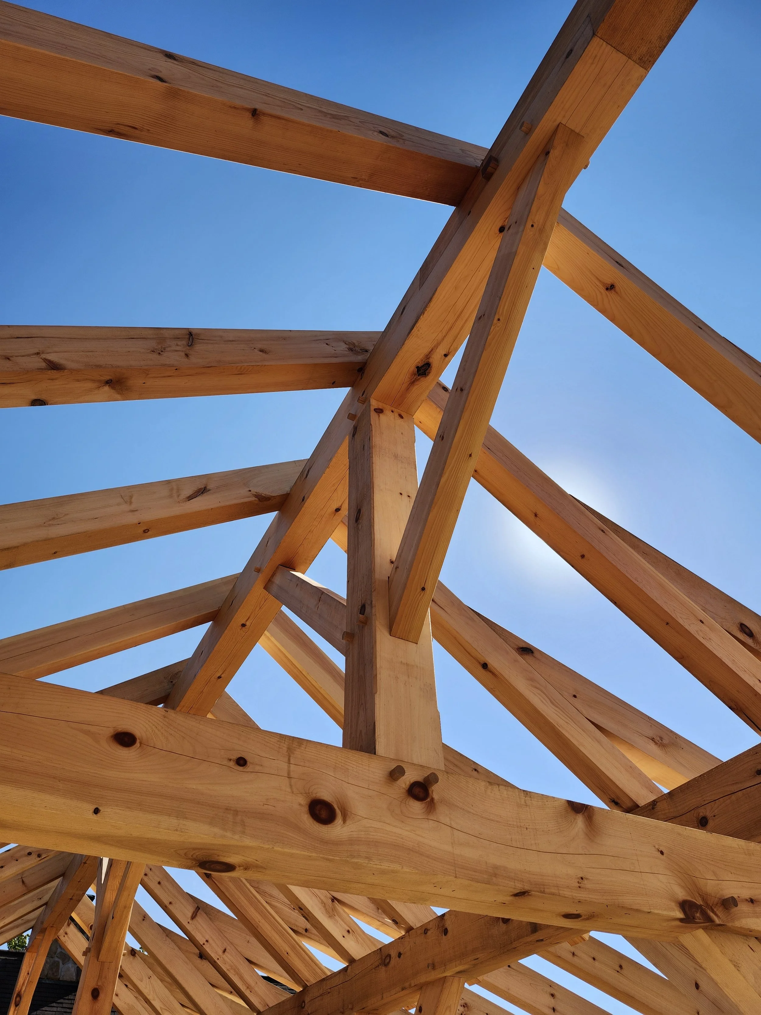 View looking up at a wooden roof frame under construction with blue sky and sunlight visible.