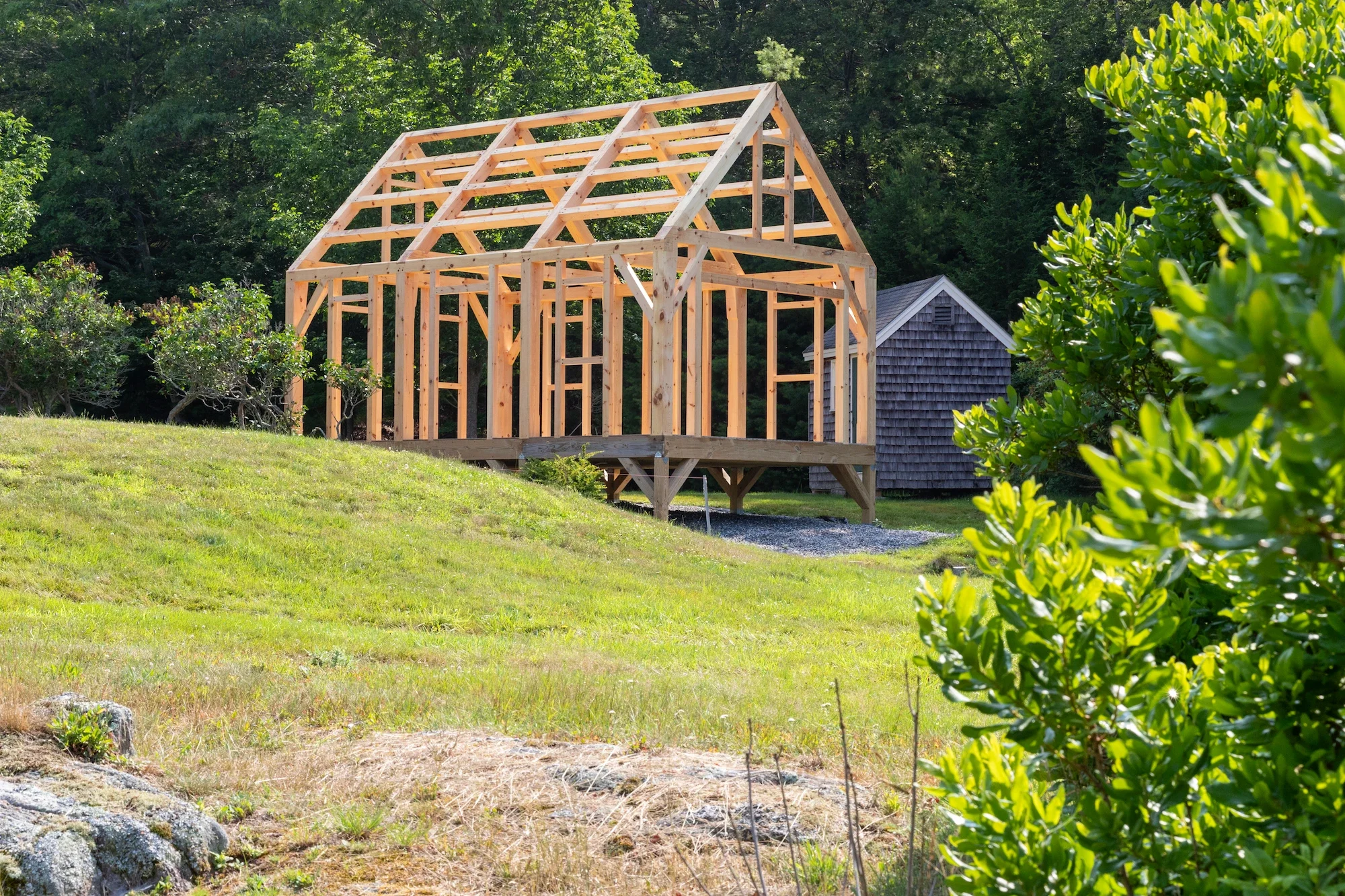 A house under construction with a wooden frame on a raised platform outdoors, surrounded by green trees and grass, with a small gray shed in the background.