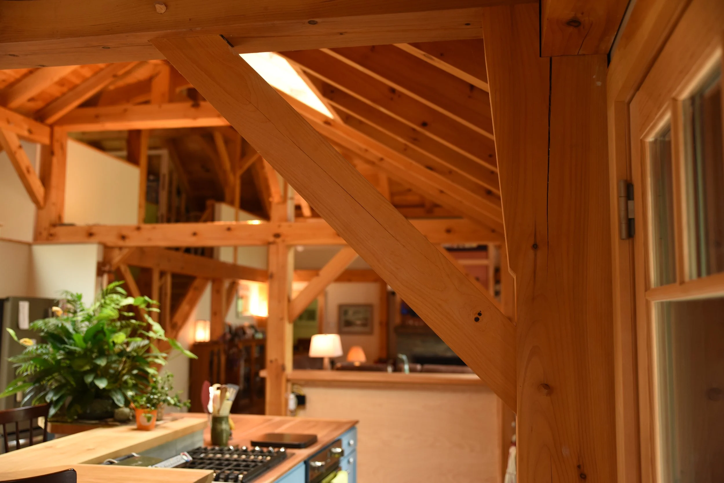 Interior view of a cozy wooden house with exposed wood beams, a stove, and a kitchen counter with a plant on it, illuminated by warm lighting.