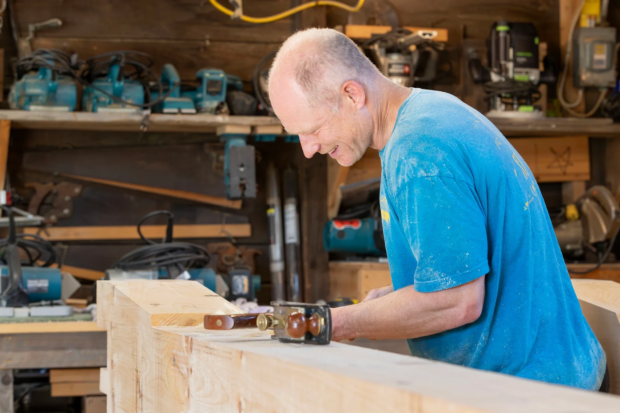 A man working on a carpentry project in a workshop, sanding a large piece of wood with a hand sander, wearing a blue shirt with sawdust on it.