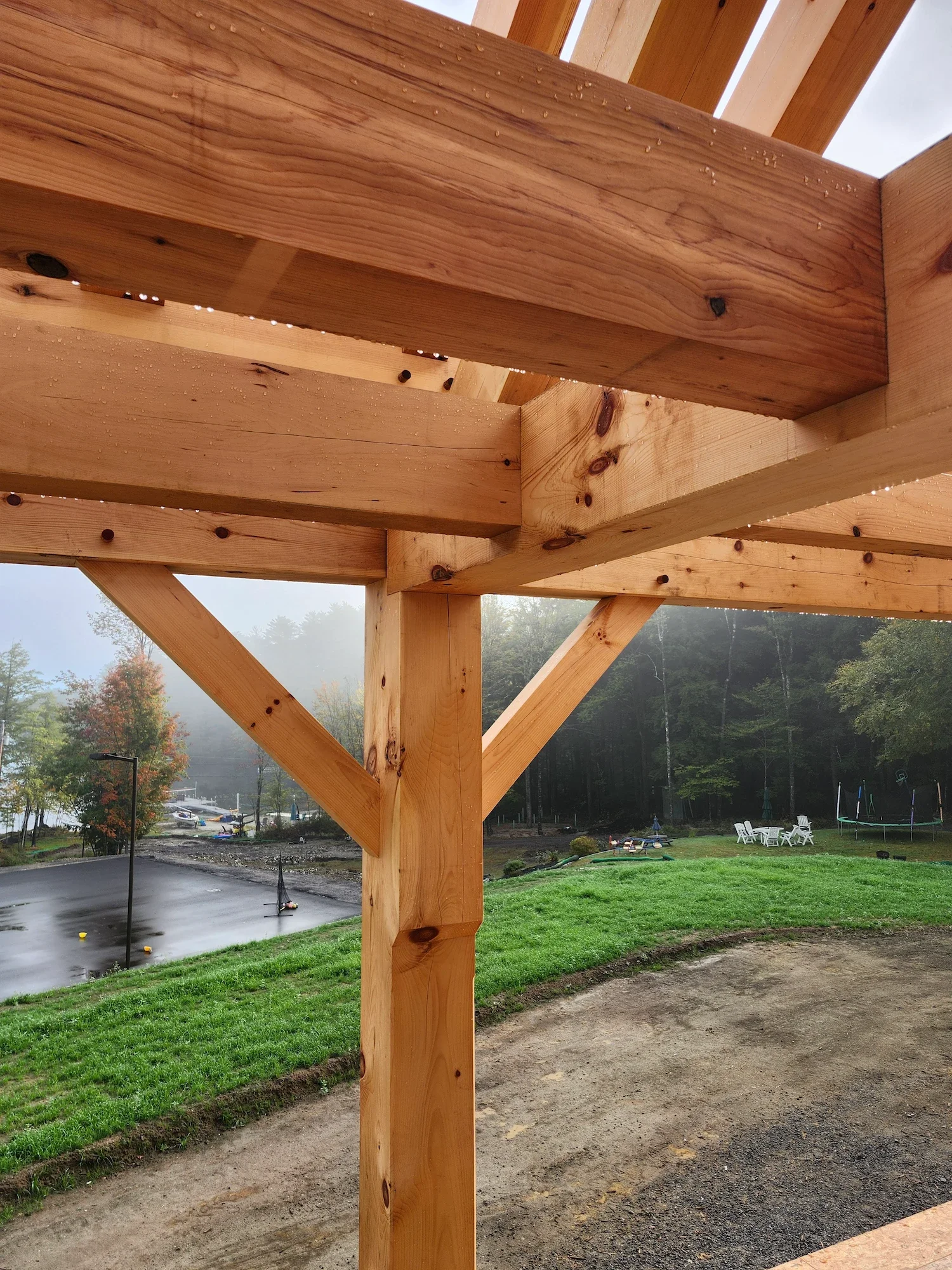 Close-up of a wooden structure under construction with visible beams and posts, outdoor area with grass, trees, and playground in the background.