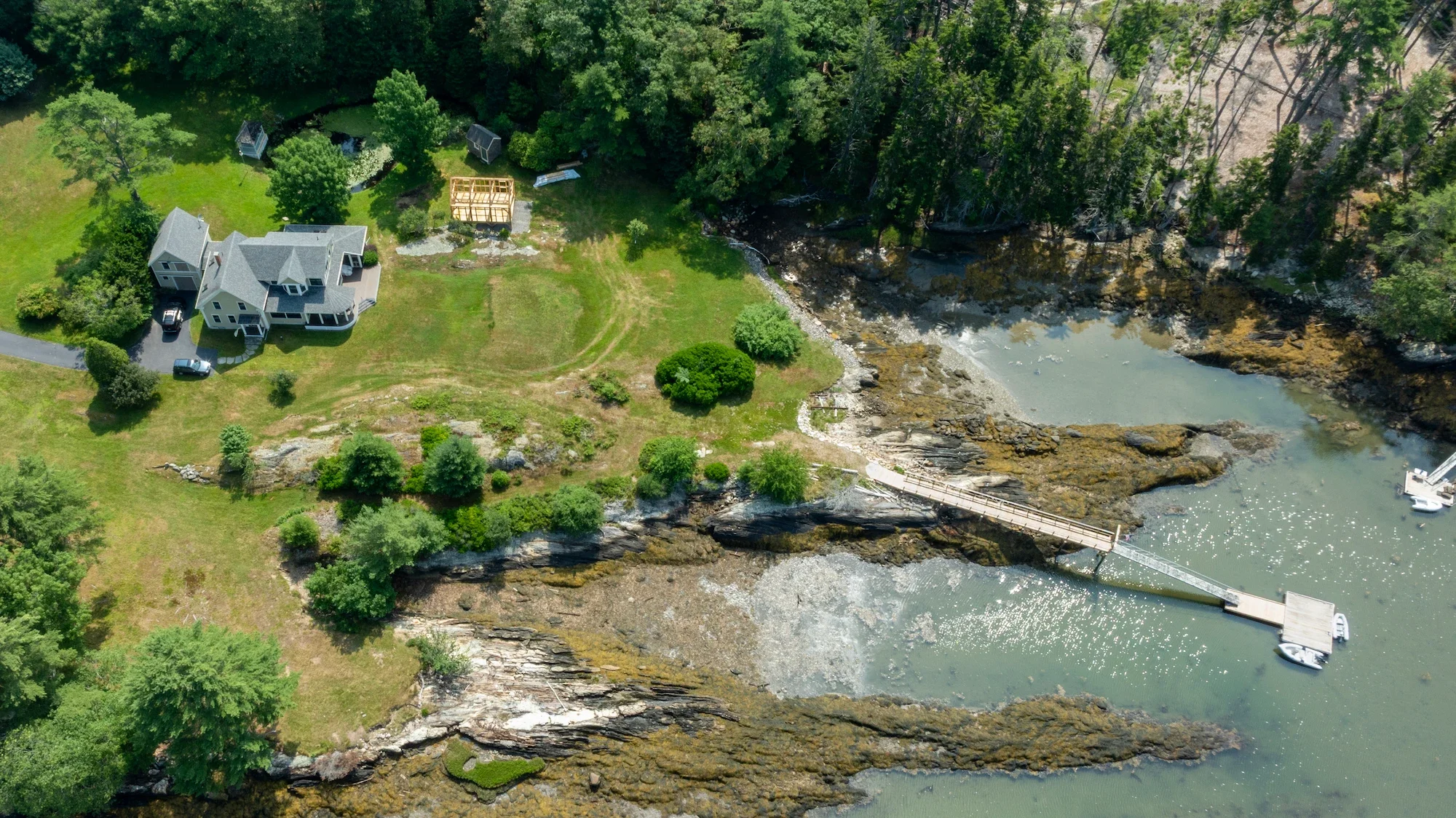 Aerial view of a lakeside property featuring a large gray house with multiple decks, a driveway, lush green lawn, and surrounding trees. There is a wooden dock extending into the water with boats moored, along with a small waterfall or rapids in the 
