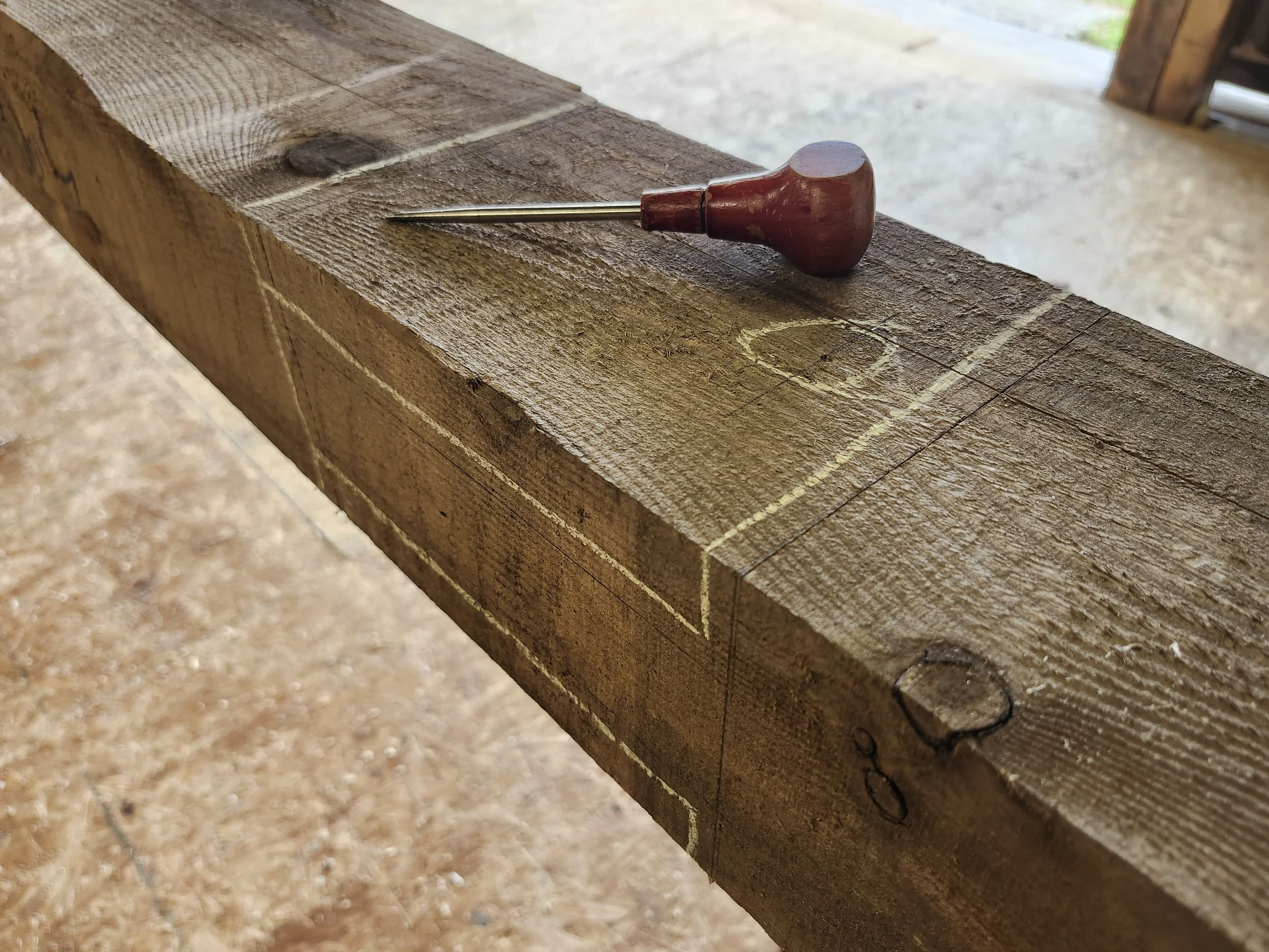 A woodworking marking gauge with a wooden handle and a metal pin, resting on a rough-cut wooden board with pencil markings and lines.