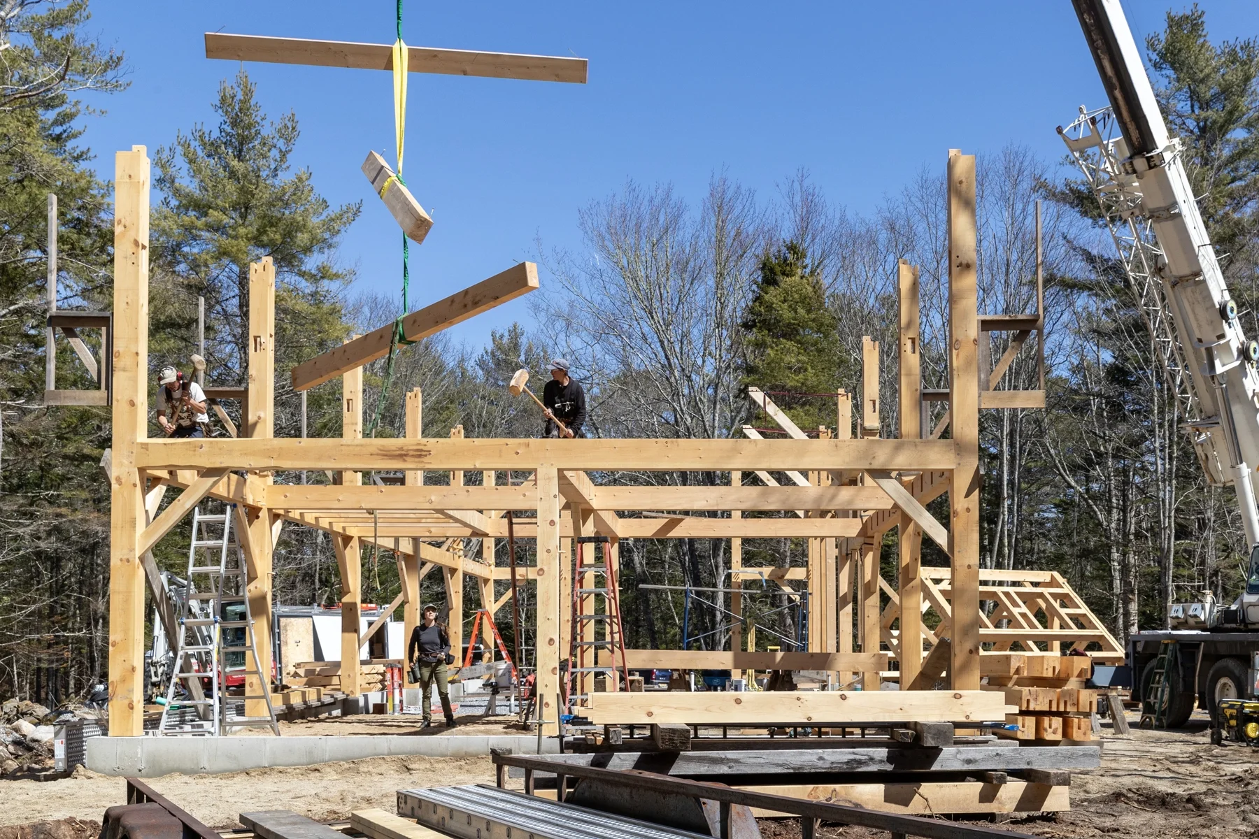 Construction workers building a wooden structure outdoors with a crane and various tools on a clear, sunny day.