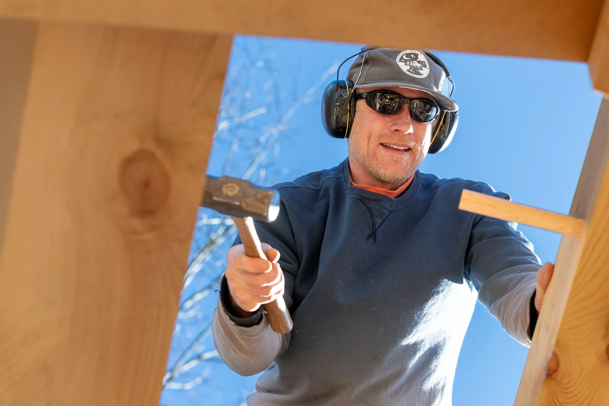 Man wearing sunglasses, a cap with a logo, and noise-canceling headphones, working on a wooden structure outdoors on a sunny day.