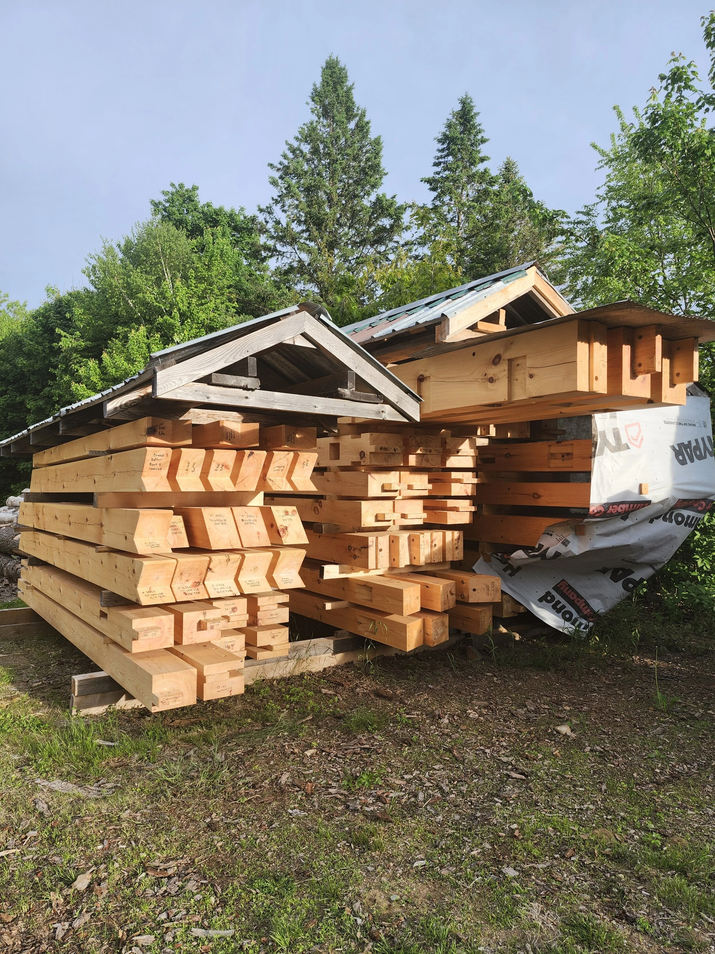 Stacks of wooden lumber with two small wooden structures, surrounded by green trees and grass, under a blue sky.