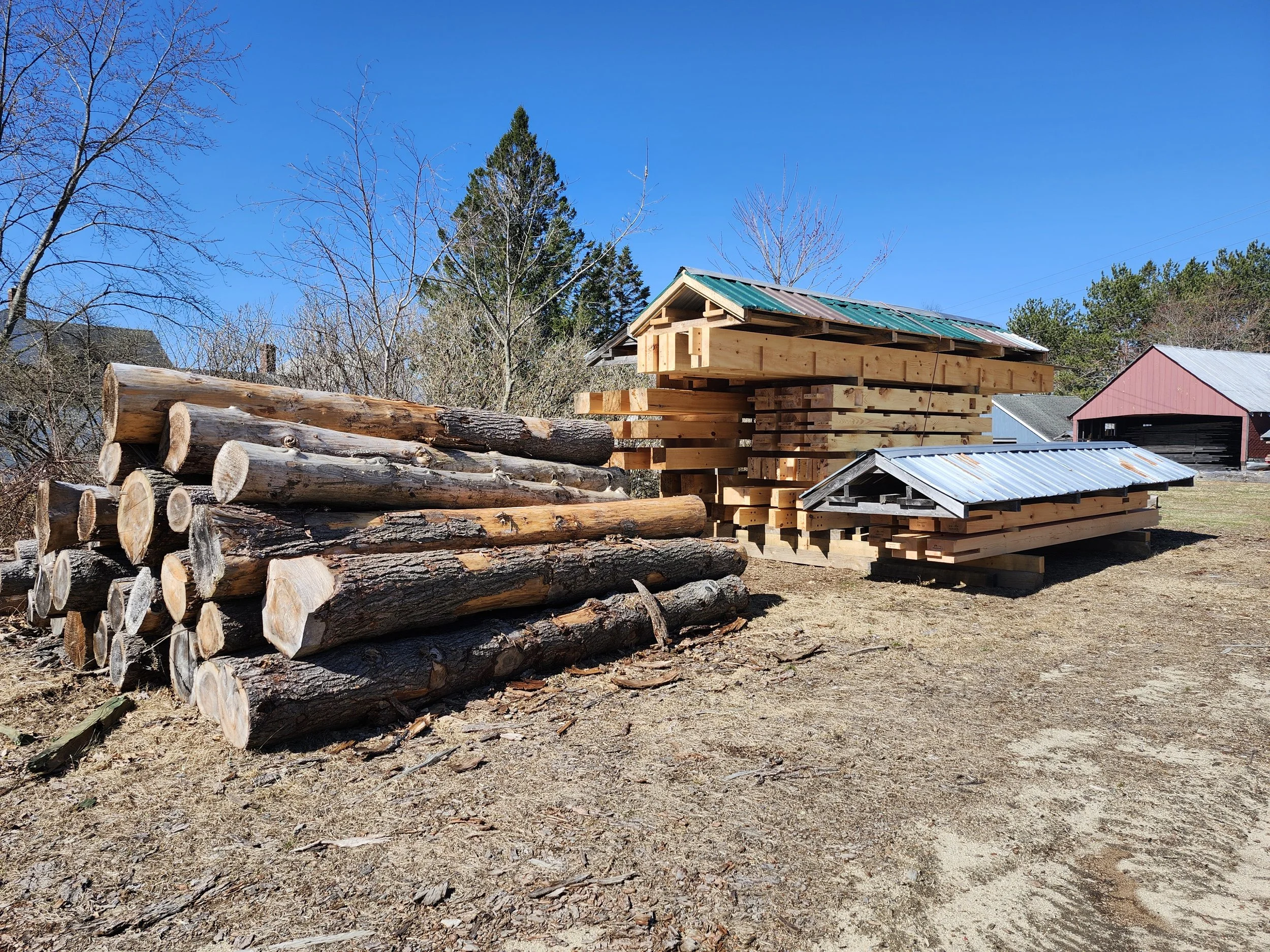 Stacks of cut logs and lumber with small sheds on a farm or construction site under a clear blue sky.