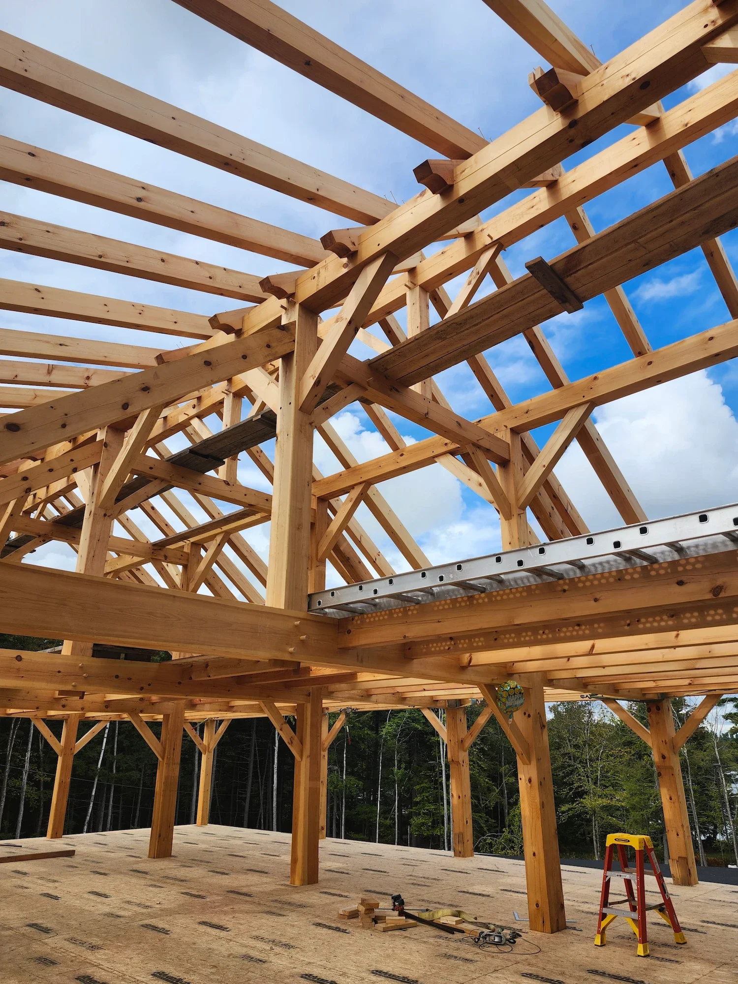 Construction site of a wooden house frame, showing beams and roof trusses, with construction tools and a step ladder on the floor.