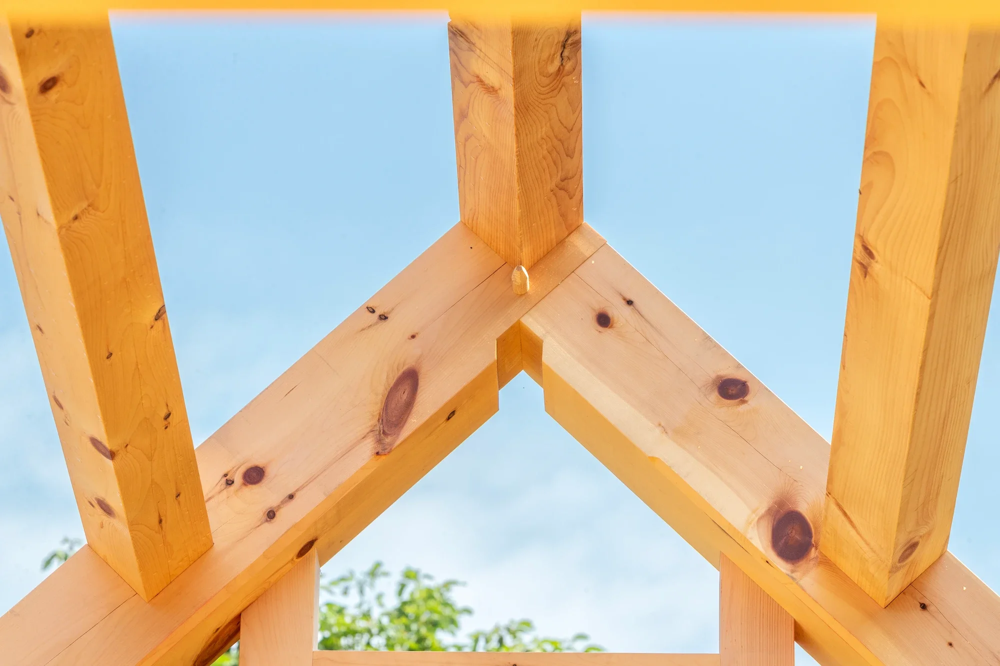 Close-up of a wooden roof truss under construction with blue sky and green trees in the background.