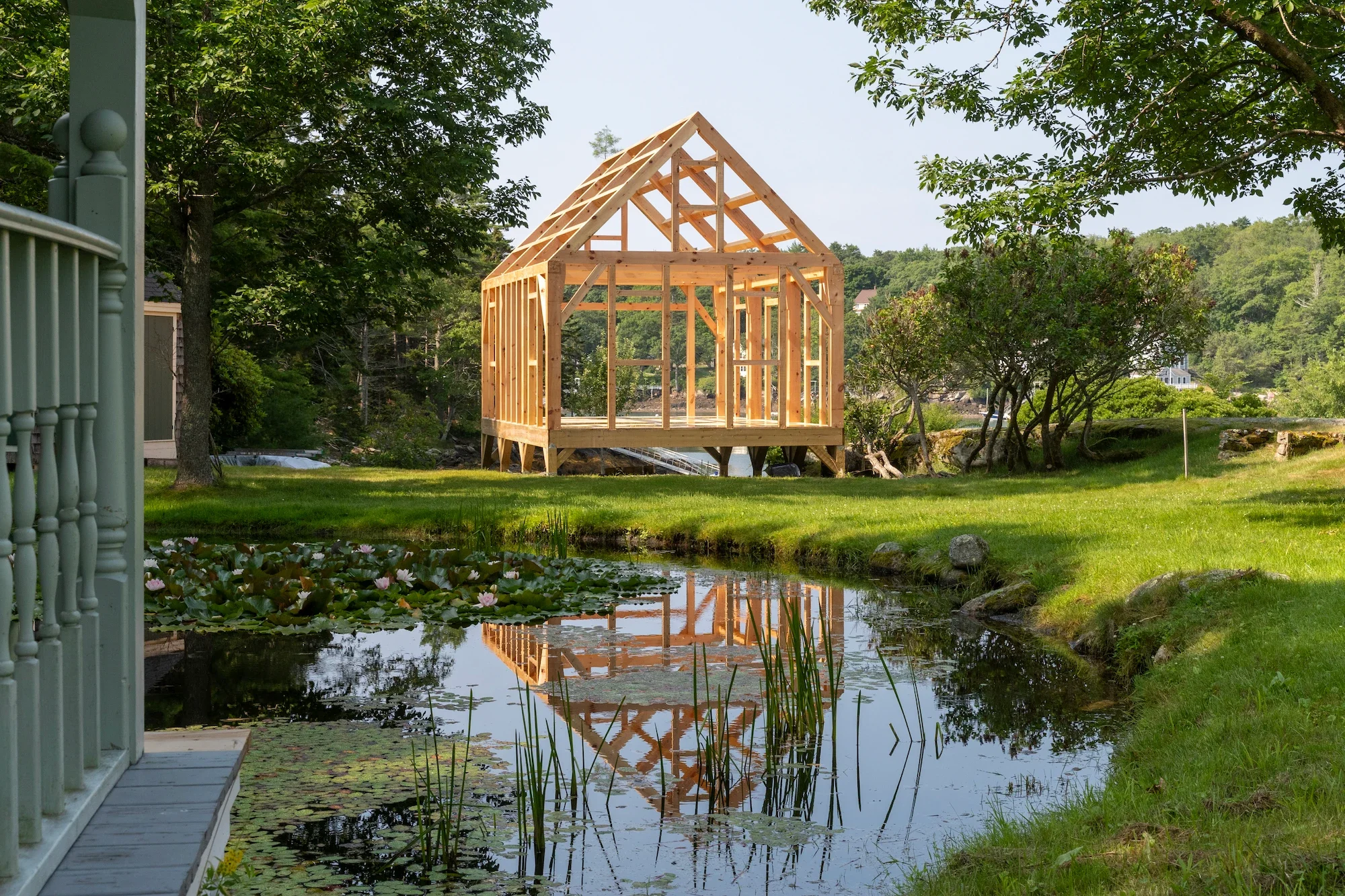A wooden house under construction near a pond with water lilies, surrounded by green trees and grass