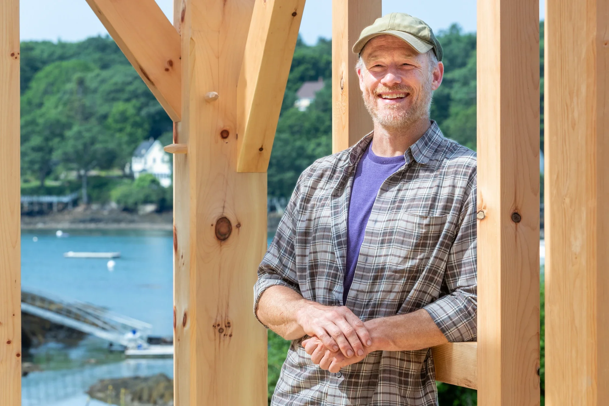 A man with a beard and a beige cap smiling while standing outdoors on a construction site, with a water and waterfront view in the background.