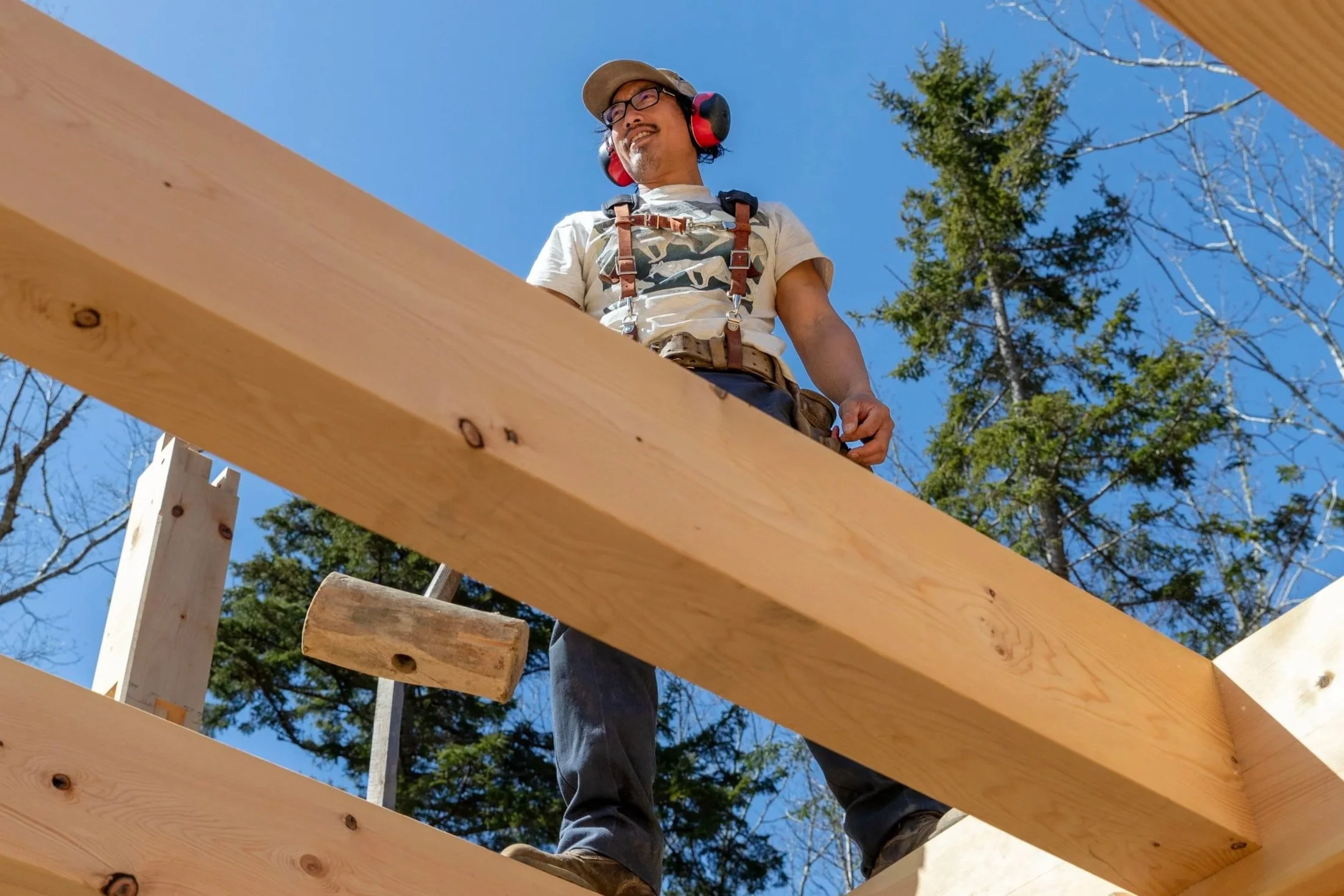 A man wearing a baseball cap, glasses, a white t-shirt, and hearing protection, standing on a wooden structure during construction, with trees and a blue sky in the background.