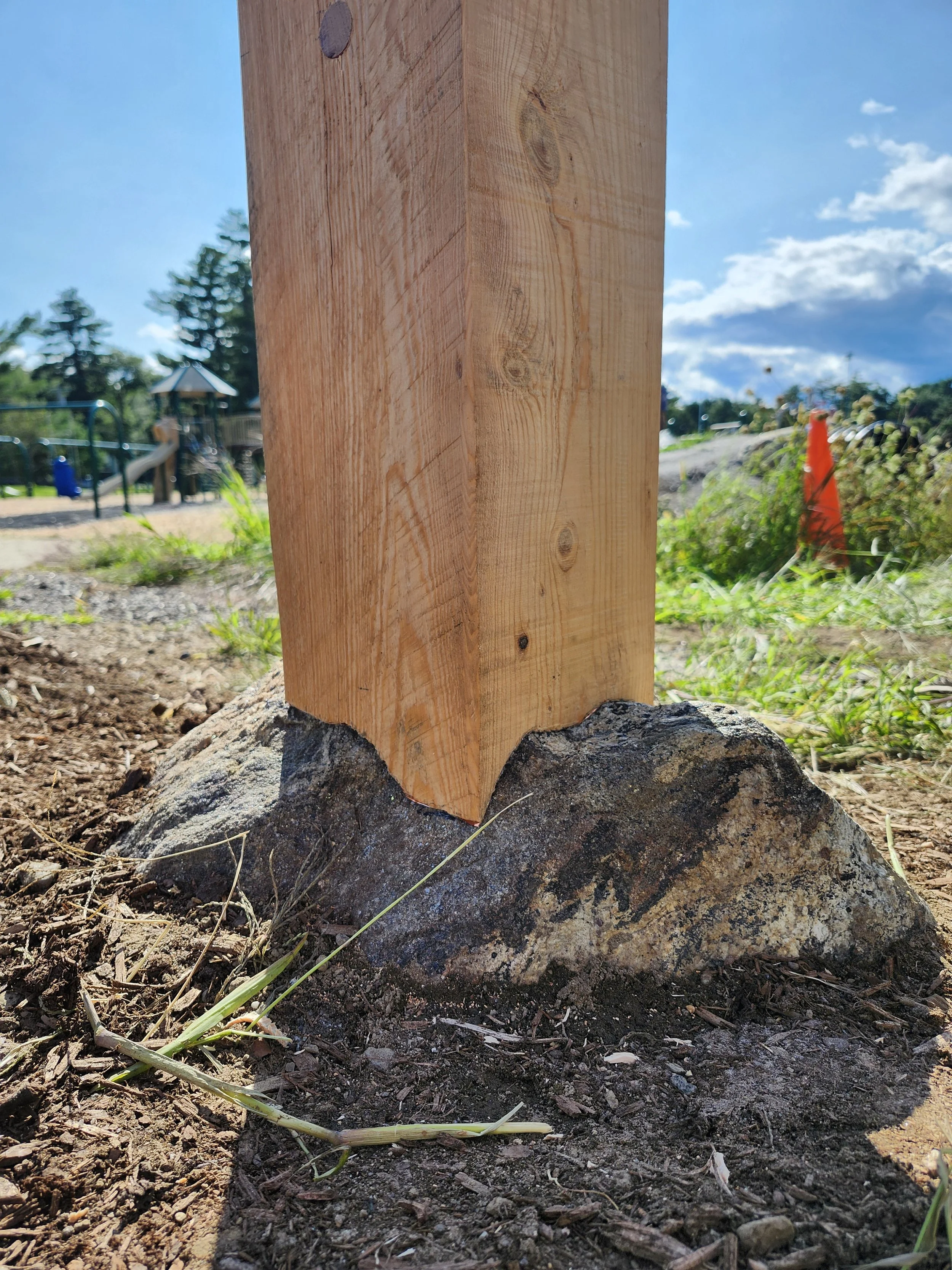 Close-up of a wooden post mounted on a large rock at a playground under a blue sky with clouds.