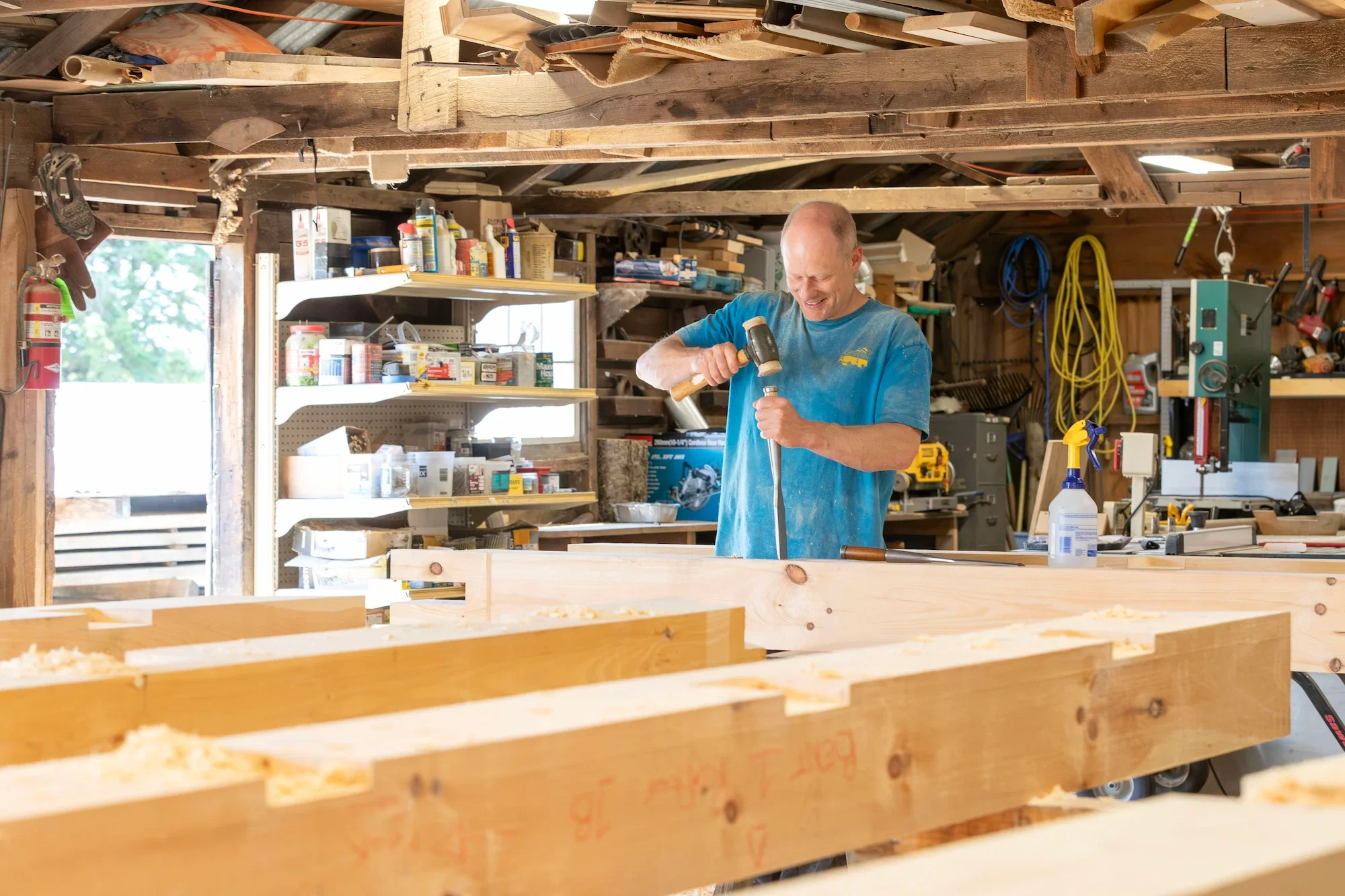 A man working in a woodworking shop, using a hammer on a large wooden plank, with woodworking tools and supplies visible in the background.