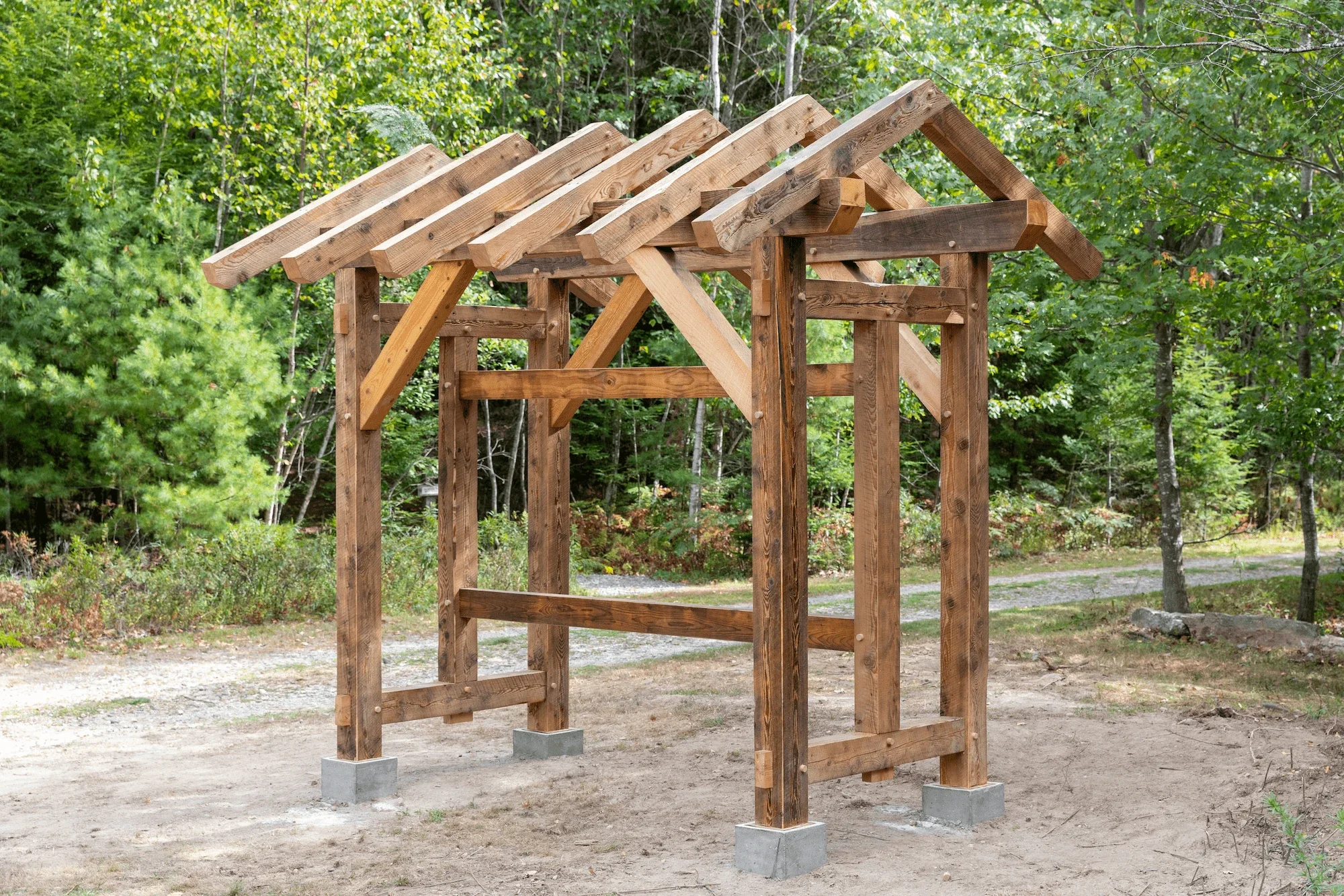 Unfinished wooden playhouse frame with a pitched roof, situated outdoors on a dirt ground surrounded by trees and greenery.