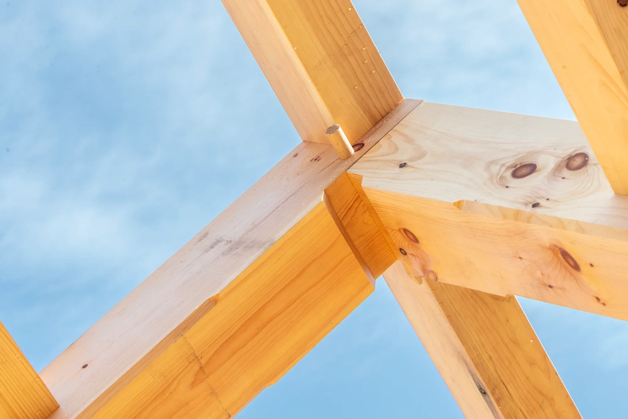 Close-up view of a wooden roof truss under construction against a blue sky, showing the joints and nails used to connect the beams.