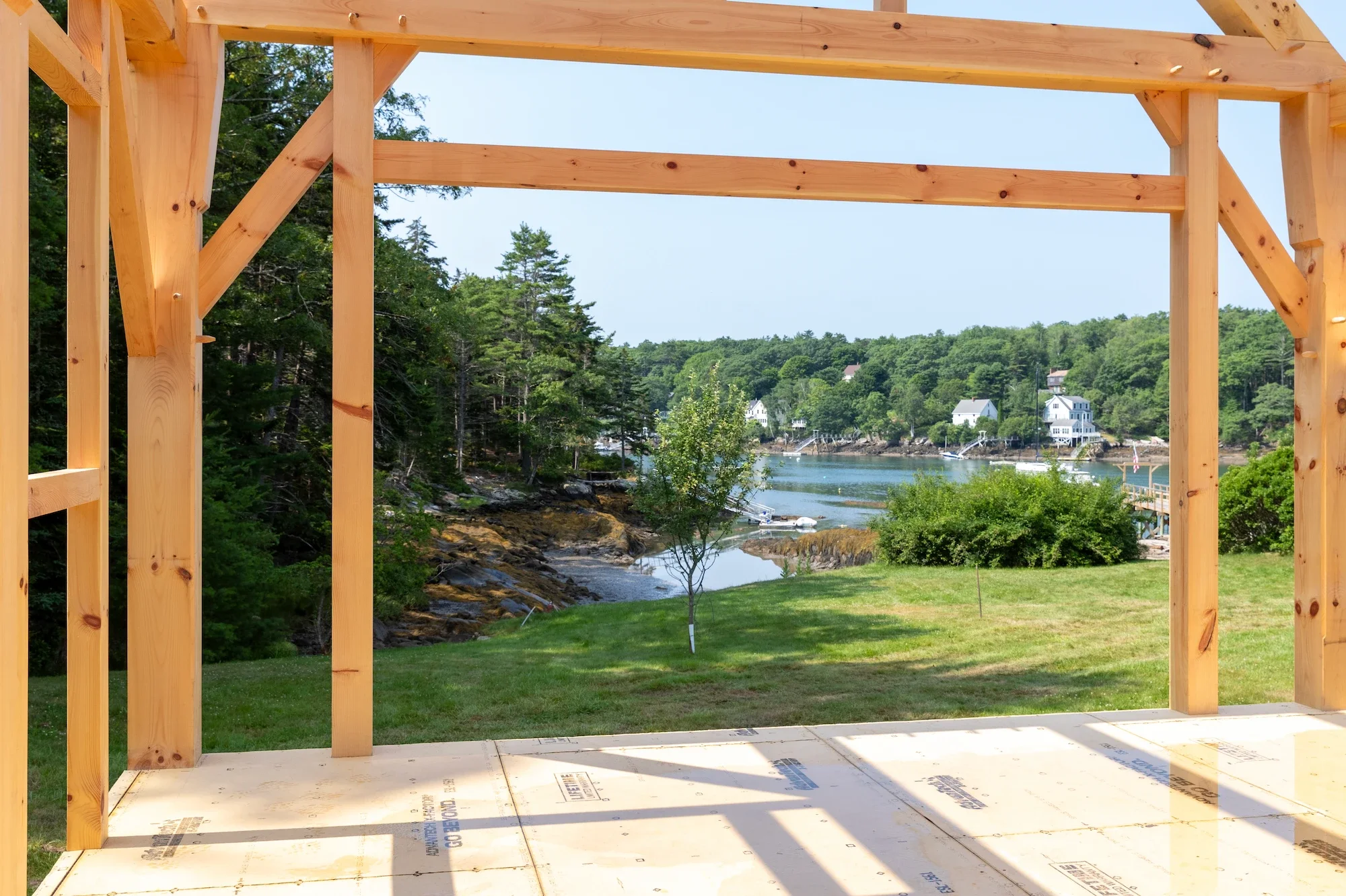 View of a waterfront scene through an unfinished wooden frame of a house under construction, with trees and houses across the water.