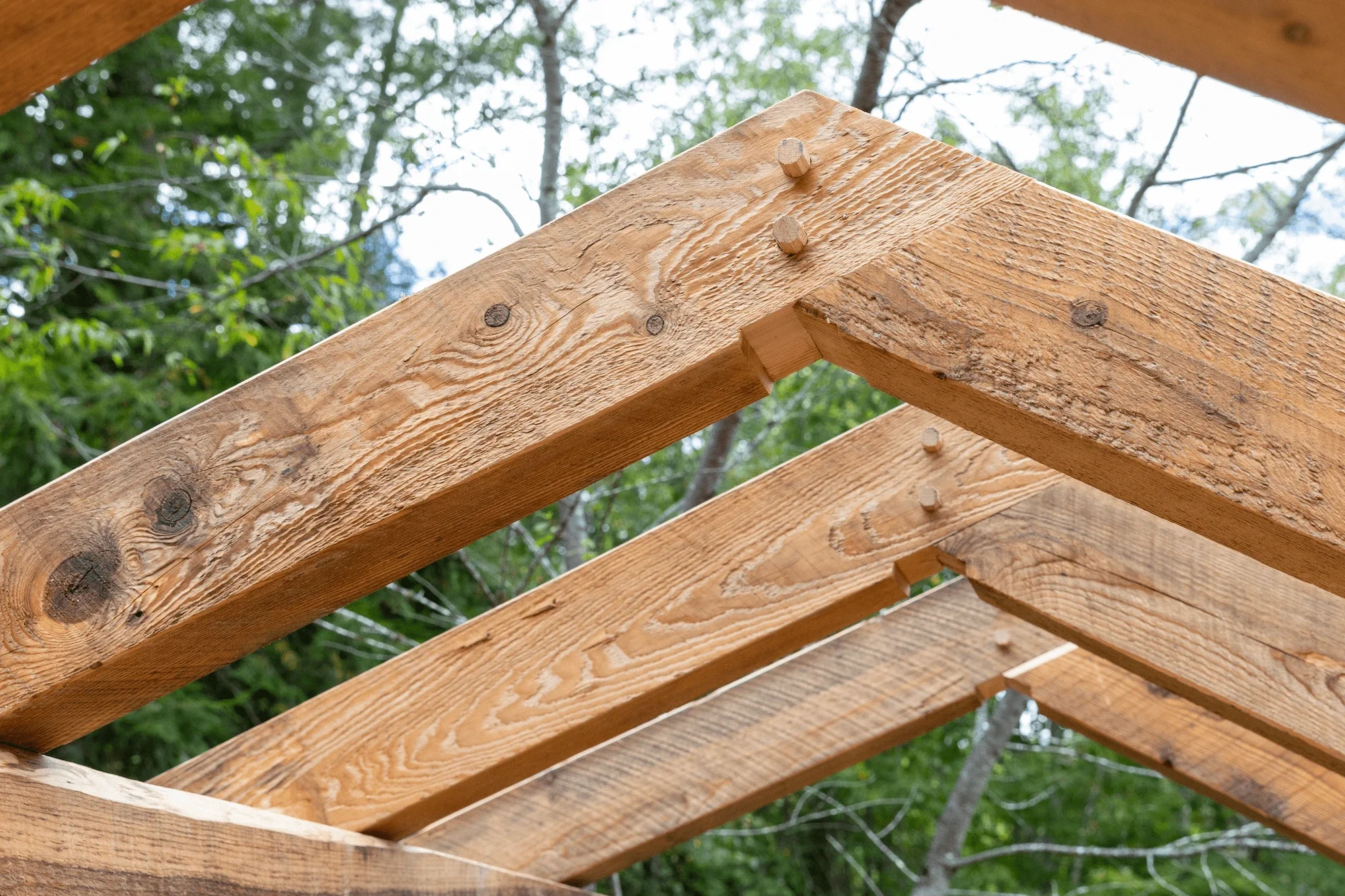 Close-up view of wooden beams and framing for a roof under construction with trees in the background.