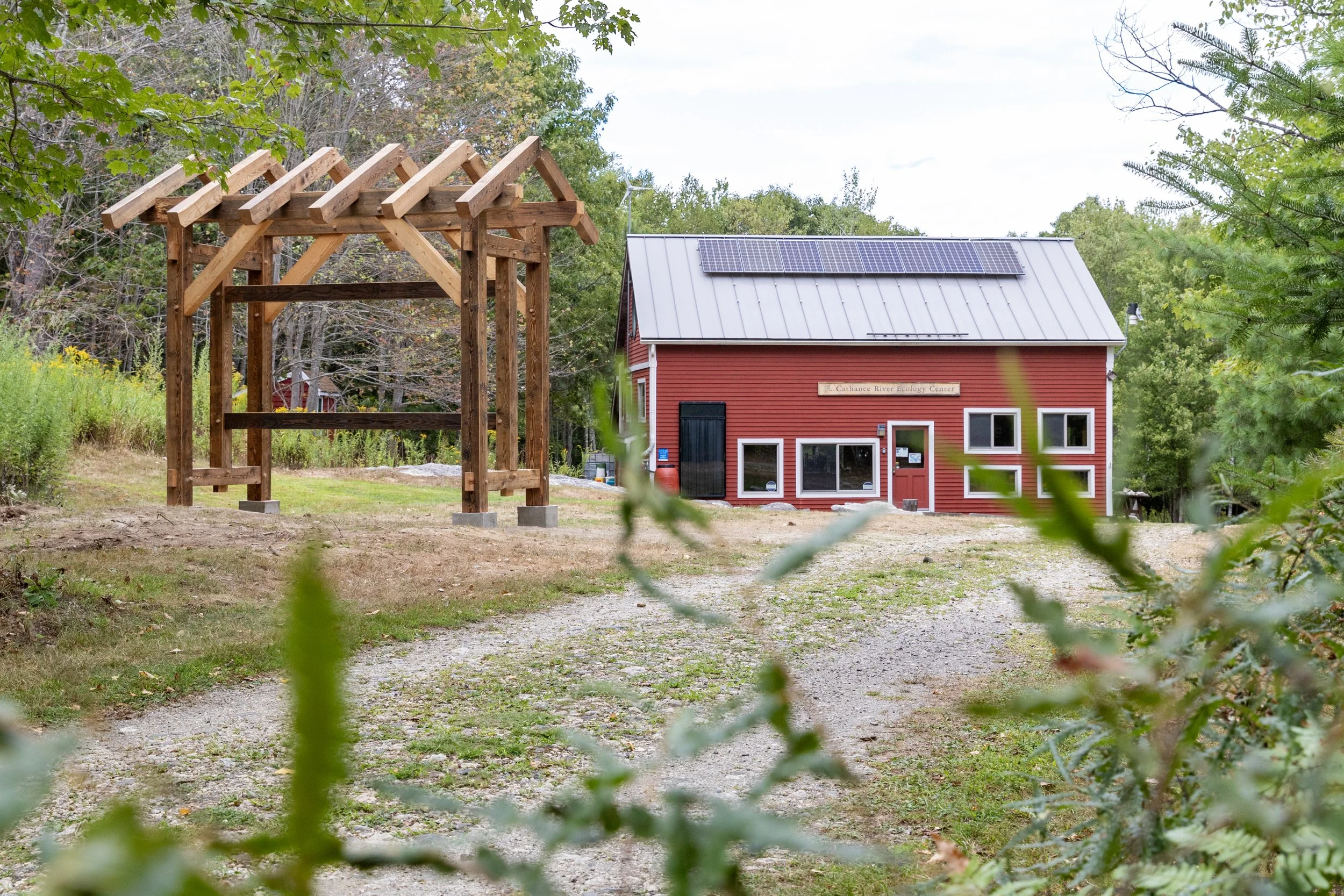 A red building with a metal roof and solar panels, labeled 'Carthance River Ecology Center,' is in the background. In the foreground, there is a wooden structure under construction, possibly a pergola, on a dirt and gravel area surrounded by green tr