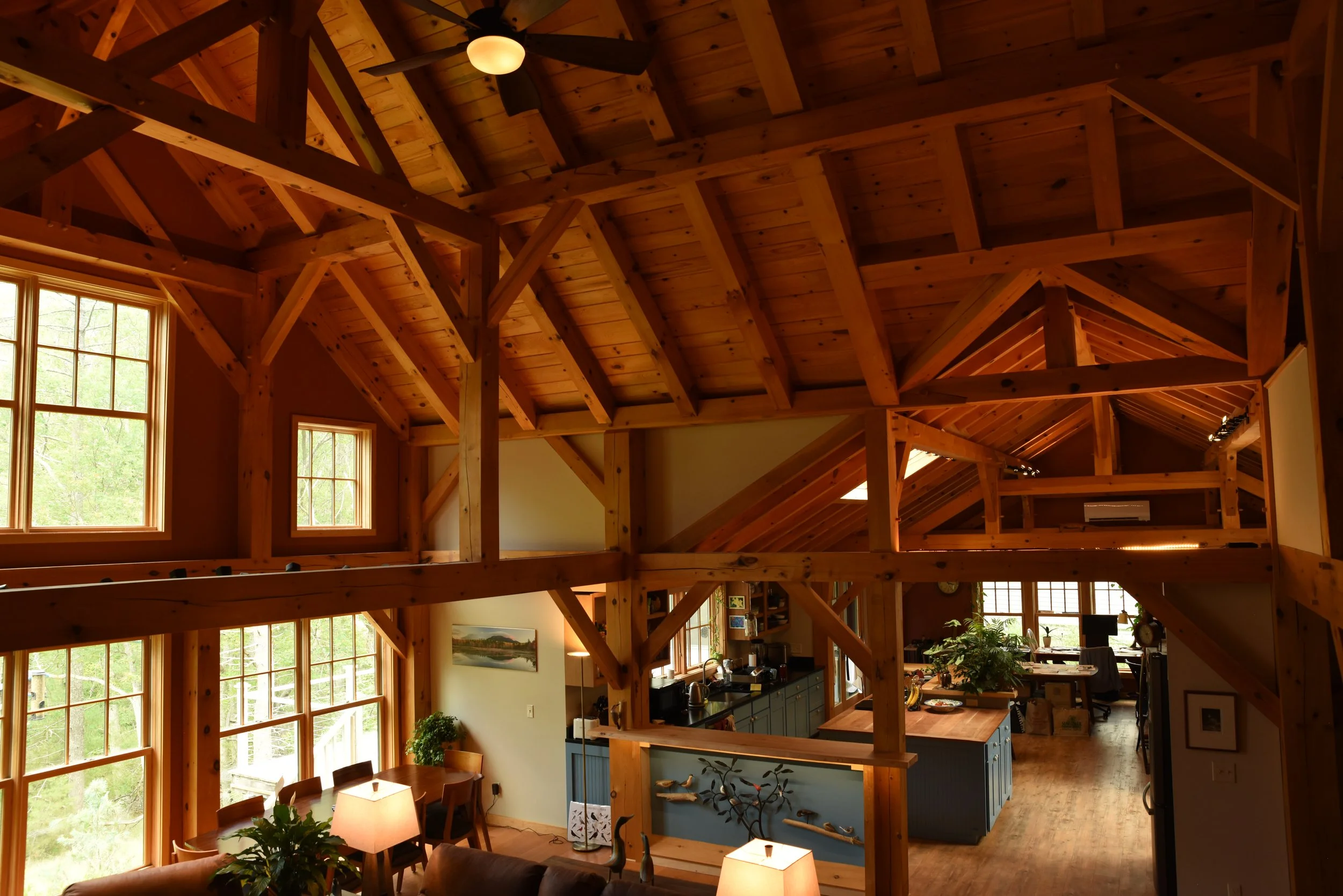 Interior view of a house with wooden beams and ceiling, large windows, dining area with a lamp, and a kitchen with blue cabinets and a small island.