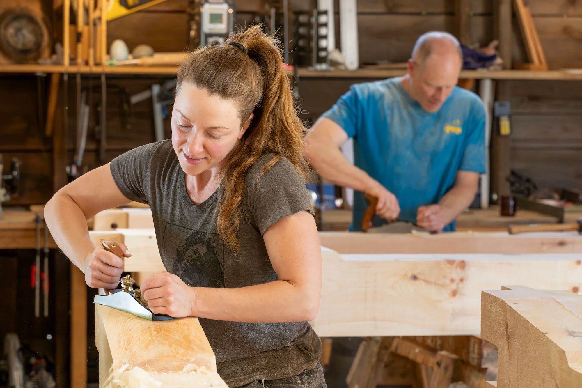 A woman and a man working on woodworking projects in a workshop. The woman in the foreground is using a hand plane on a piece of wood, and the man in the background is sanding or shaping a piece of wood.