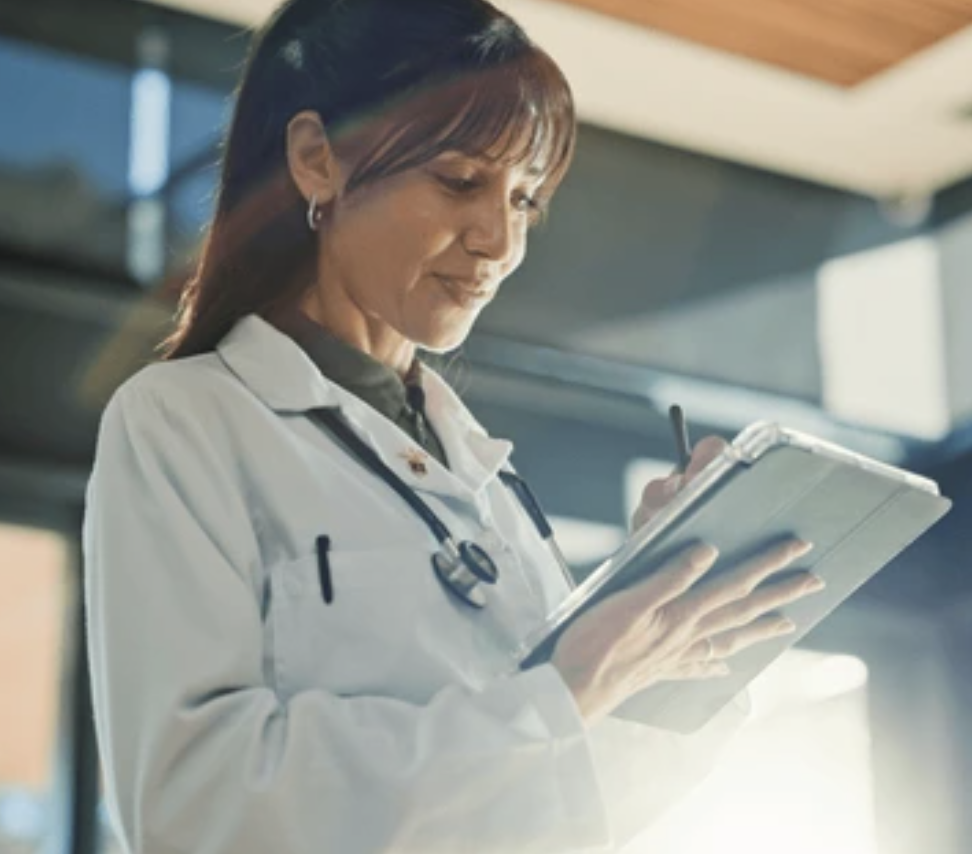 A female healthcare professional in a white coat with a stethoscope around her neck, writing on a clipboard inside a medical facility.