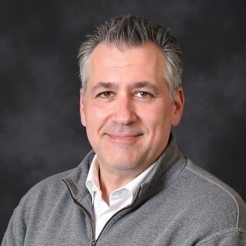 A man with short, gray hair wearing a light-colored hoodie standing in front of a dark wall.