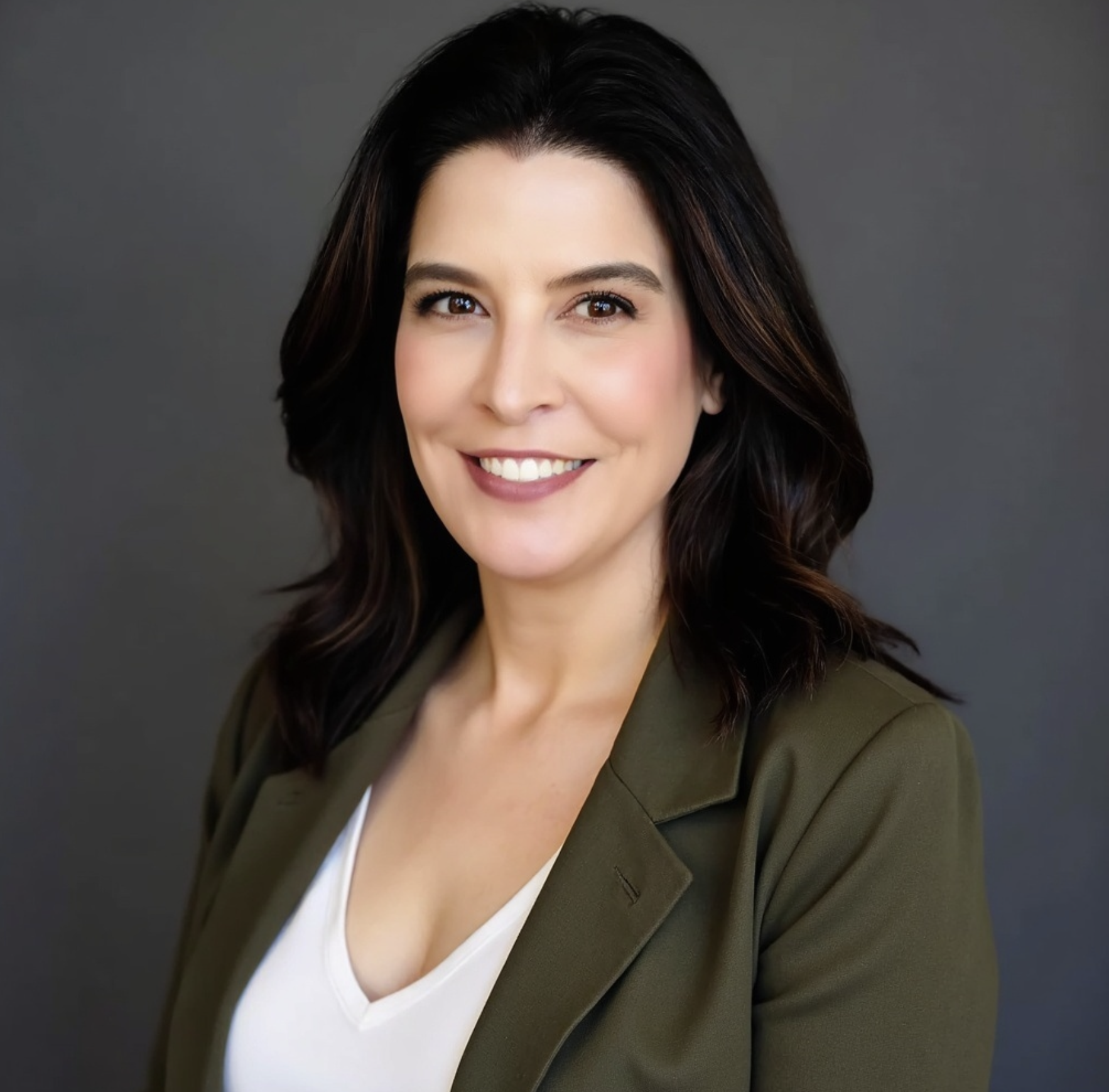 A woman smiling with black hair and earrings, wearing a dark top and jacket, in a well-lit indoor setting.