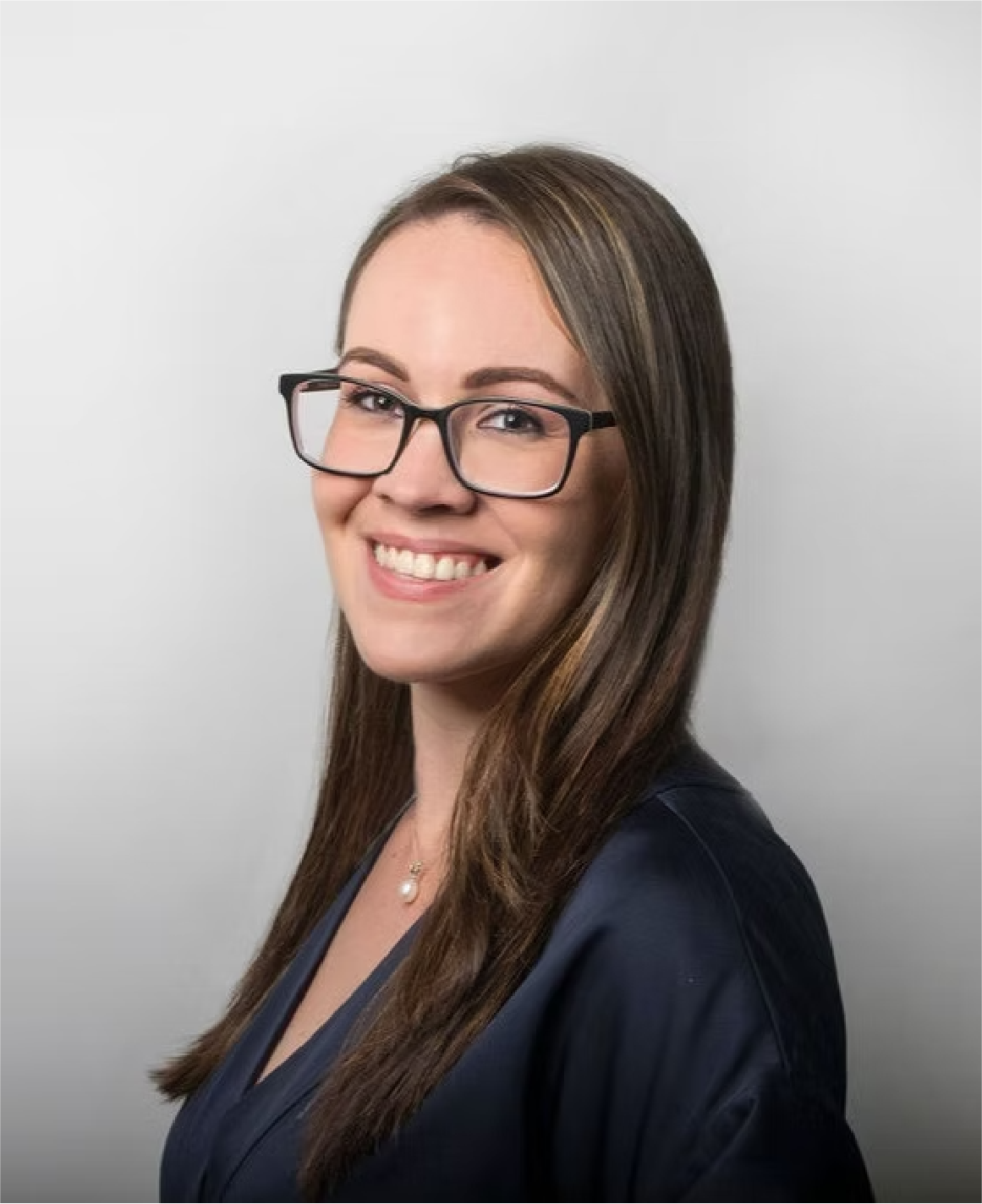 A woman with long brown hair, wearing glasses, a navy blouse, and a pearl necklace, smiling against a plain light gray background.