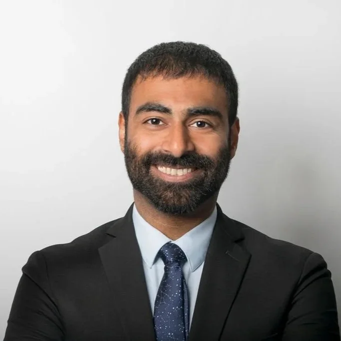 A man with dark hair and a beard wearing a black suit, light blue shirt, and a navy blue tie, smiling against a light gray background.