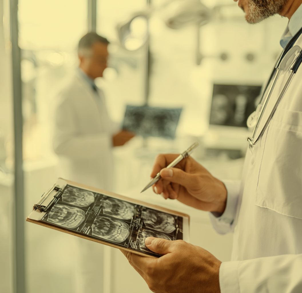 Close-up of a healthcare professional reviewing dental X-rays on a clipboard, with another medical professional blurred in the background in a clinical setting.