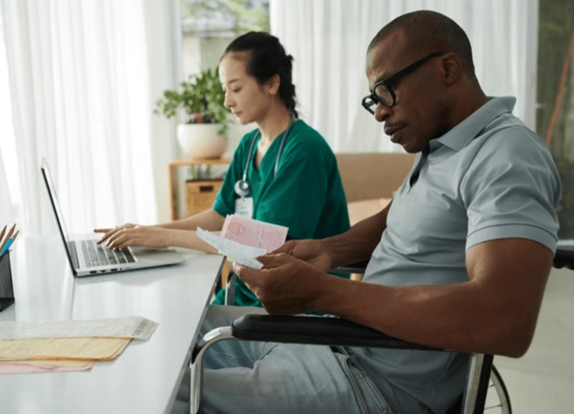 A male patient in a wheelchair reading papers and a female nurse working on a laptop in a well-lit room with curtains and a plant in the background.