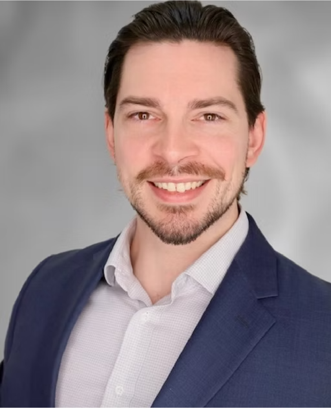 A young man with brown hair, a beard, and a mustache smiling in a professional headshot. He's wearing a white shirt and a dark blue blazer, with a plain grey background.