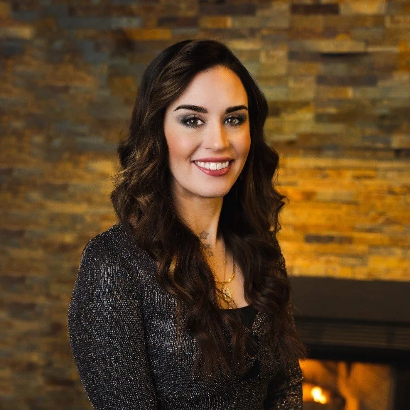 A woman with long, wavy dark hair, wearing a dark, shimmery top, smiling, with a brick wall background and a fireplace.