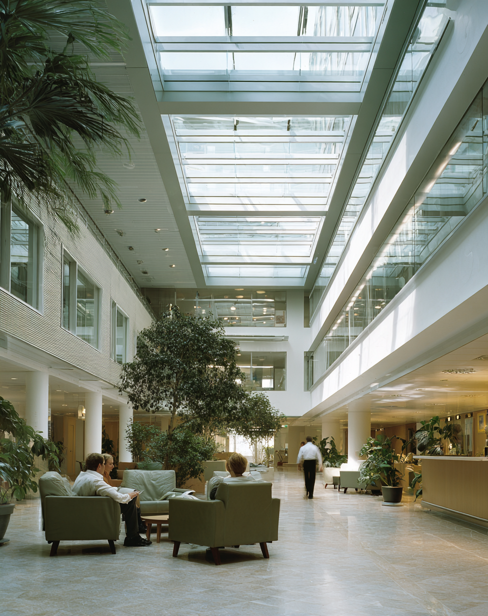 Interior of a modern office lobby with large glass ceiling, green plants, and seating area where people are relaxing and talking.