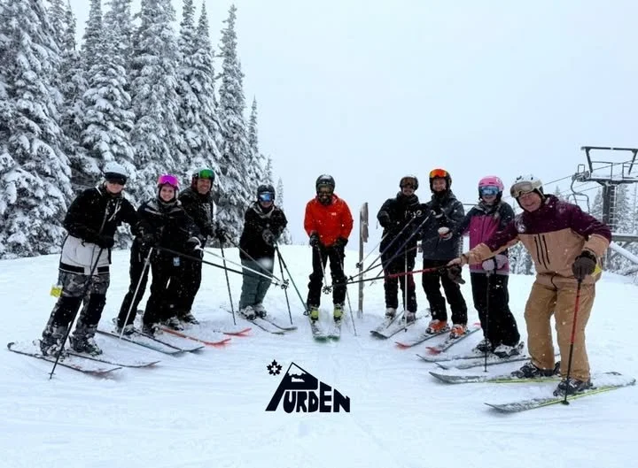 Group of nine people in skiing gear and helmets standing on snow-covered mountain slope with snow-covered trees in background, posing with skis and ski poles.