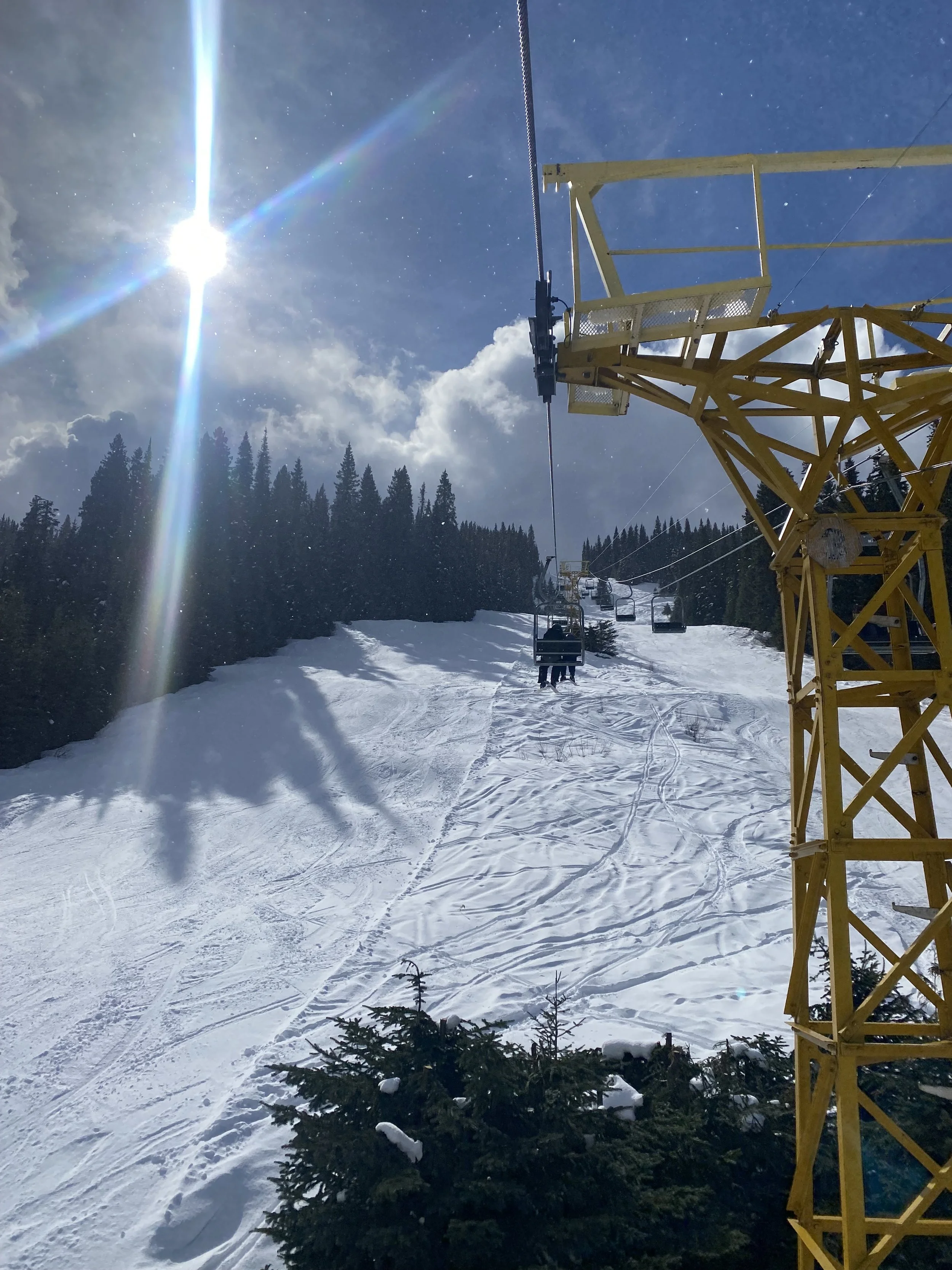Snow-covered ski slope with ski lift chairs moving up the mountain, tall evergreen trees on the side, bright sun shining in a partly cloudy sky, and a yellow ski lift tower in the foreground.