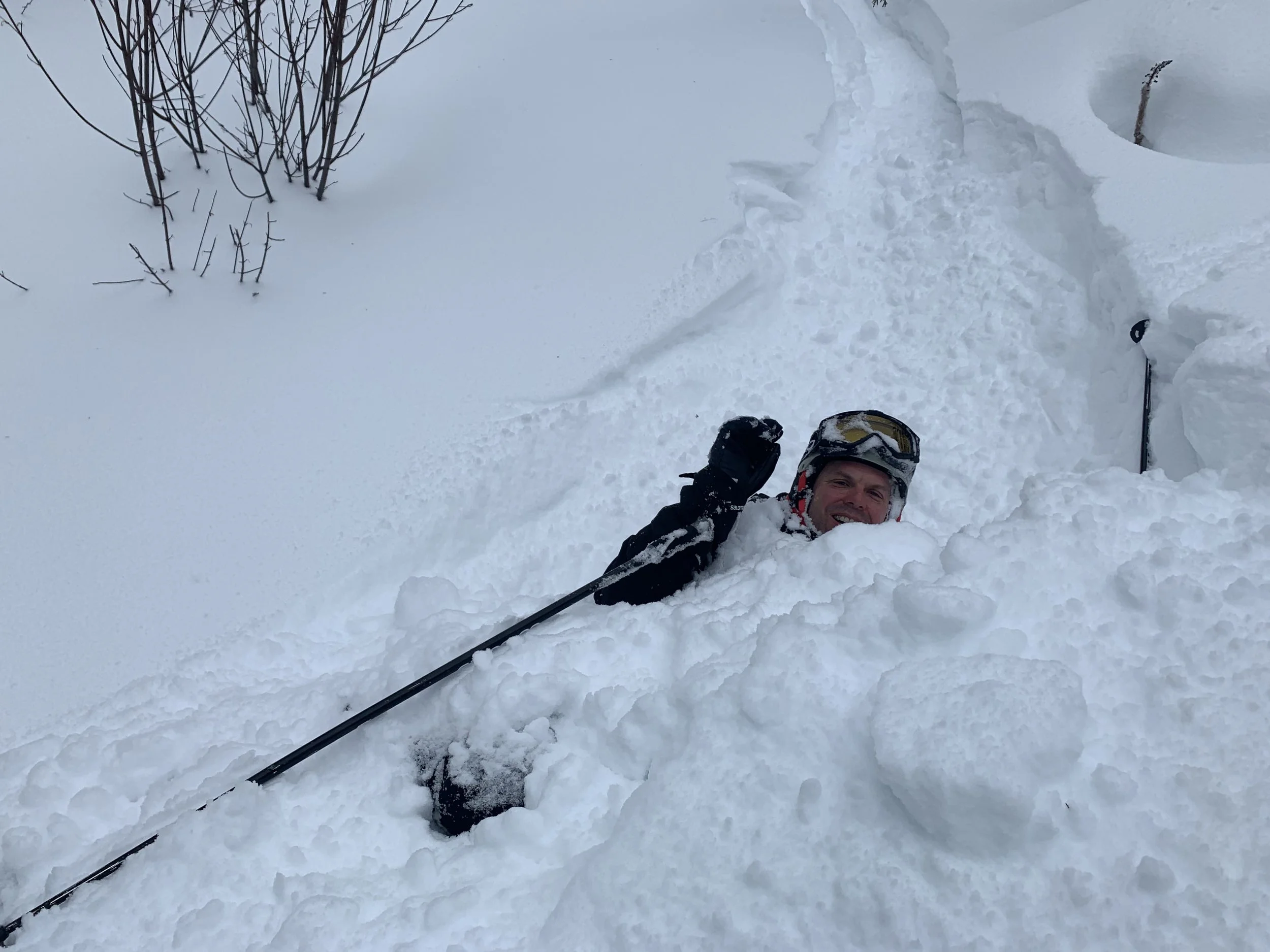 A man caught in deep snow, smiling, with a pair of ski poles and wearing a helmet, in a snowy outdoor setting with some sparse trees in the background.