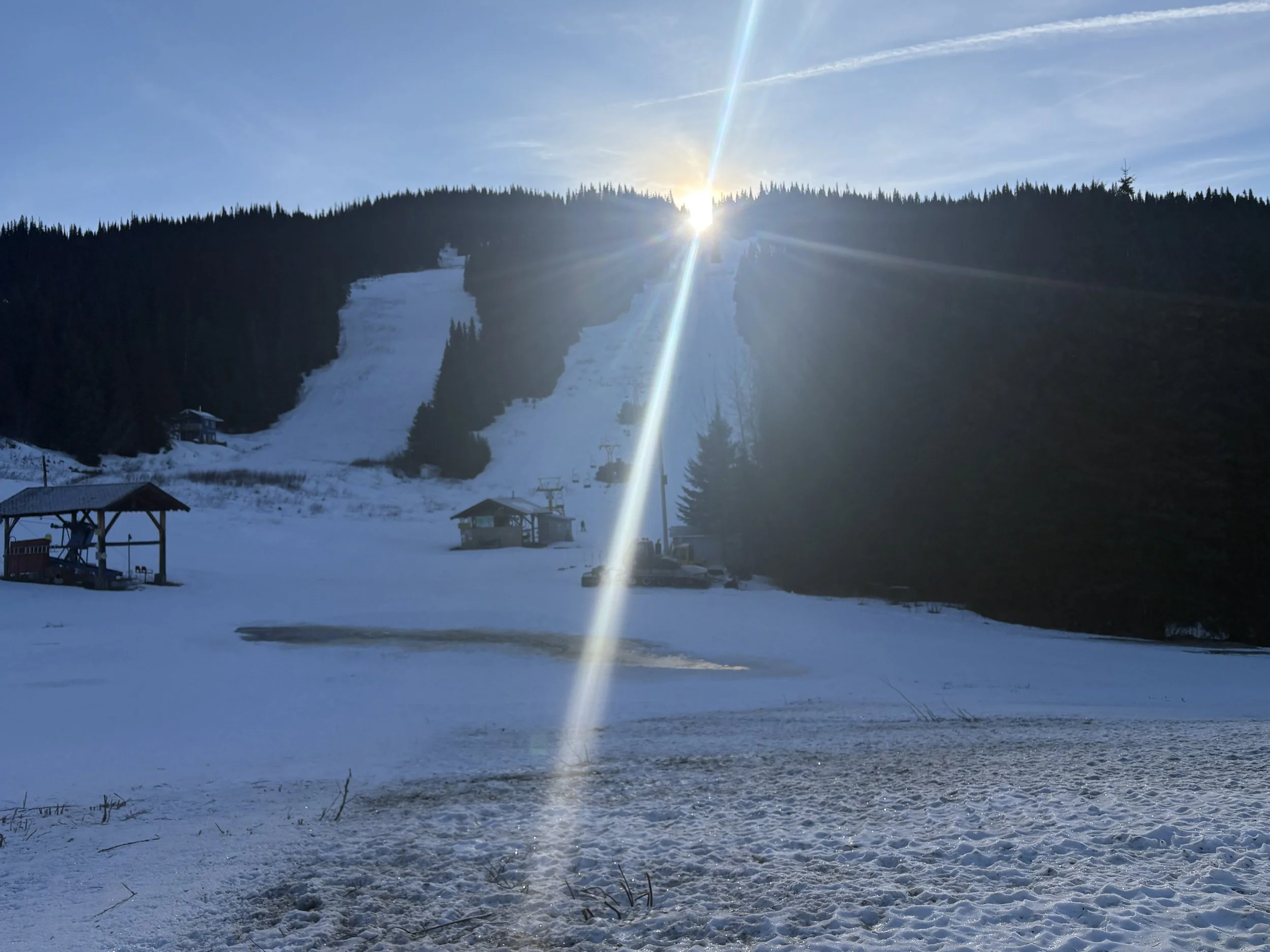 Ski resort area with snow-covered ground, ski lifts, and a ski slope on a hill beneath a bright sun in a clear sky.